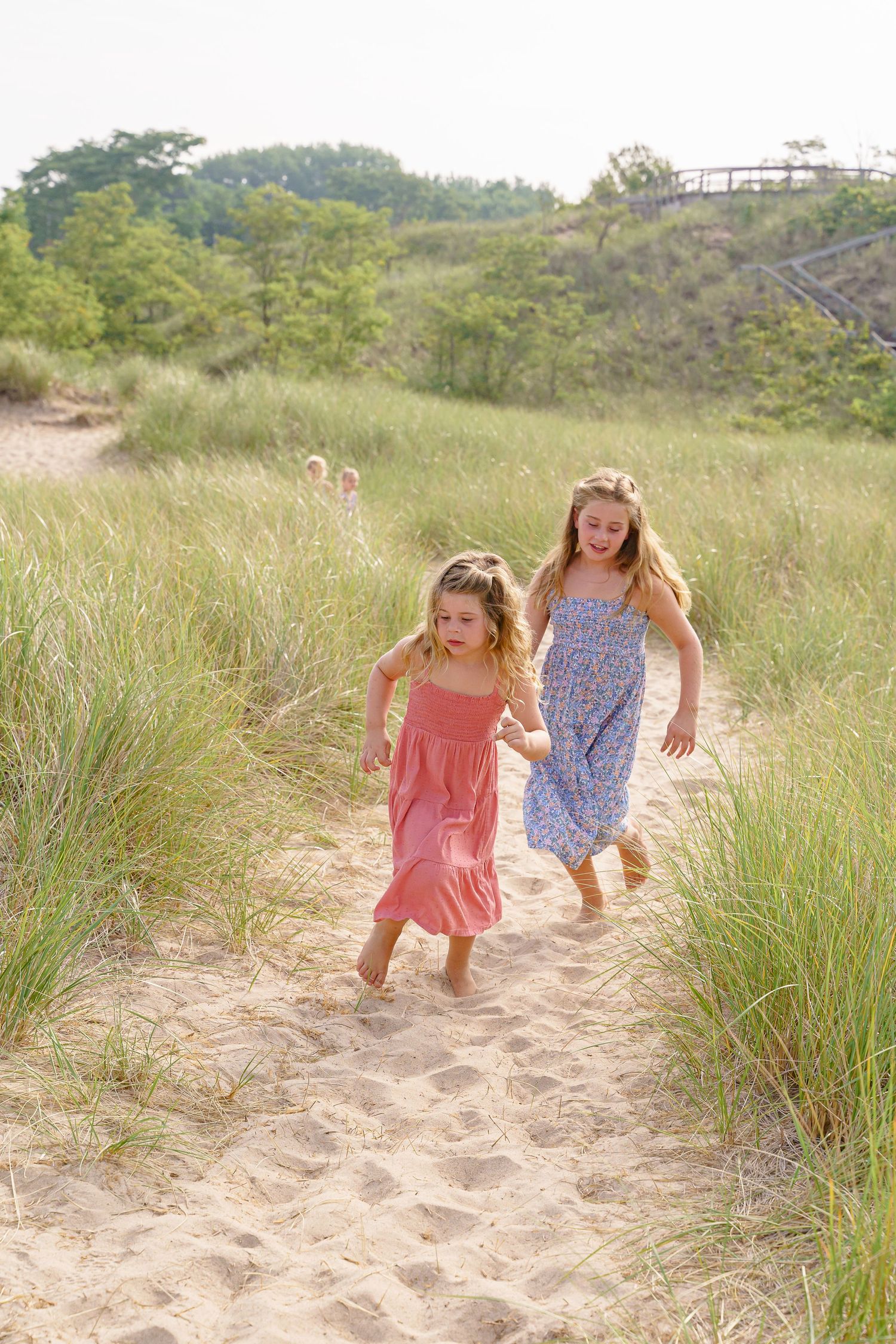 Two children in summer dresses run playfully through sandy beach dunes covered in grass.
