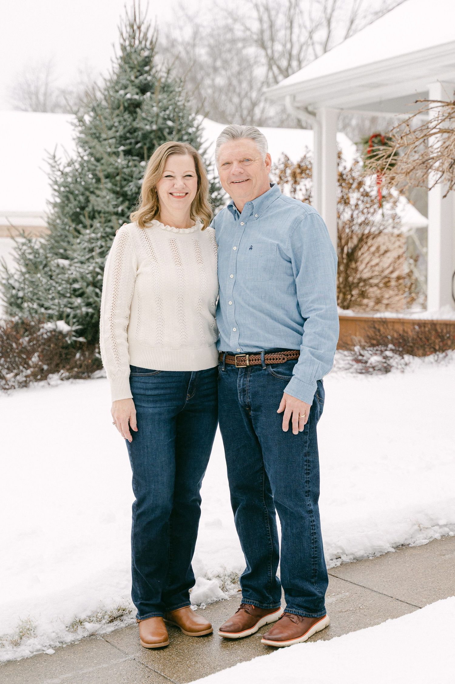 A couple stands together outdoors in winter wearing blue jeans and casual attire against a snowy white backdrop.