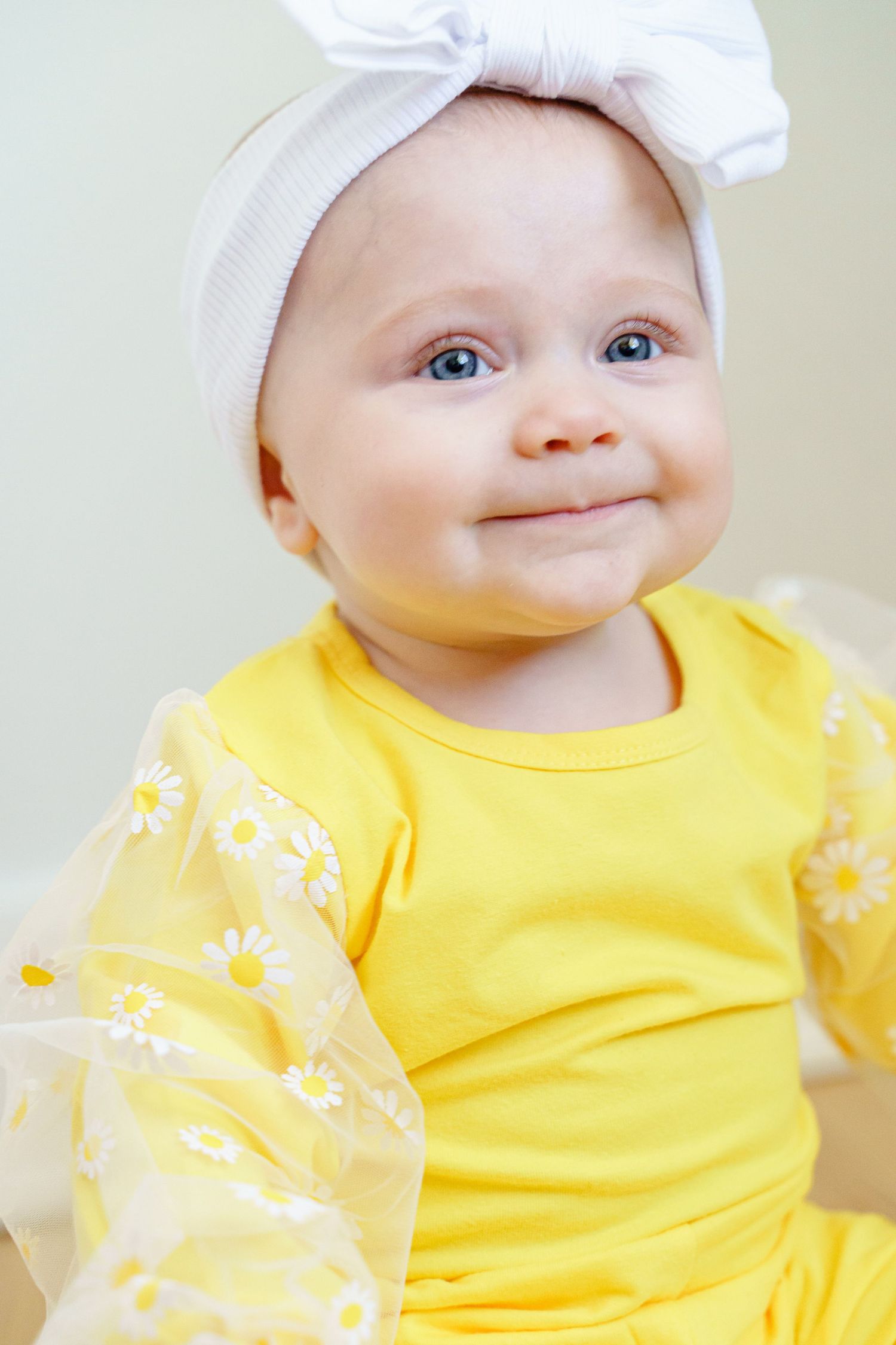 Baby in yellow outfit and white headband shows a cheerful expression while posing for a portrait.