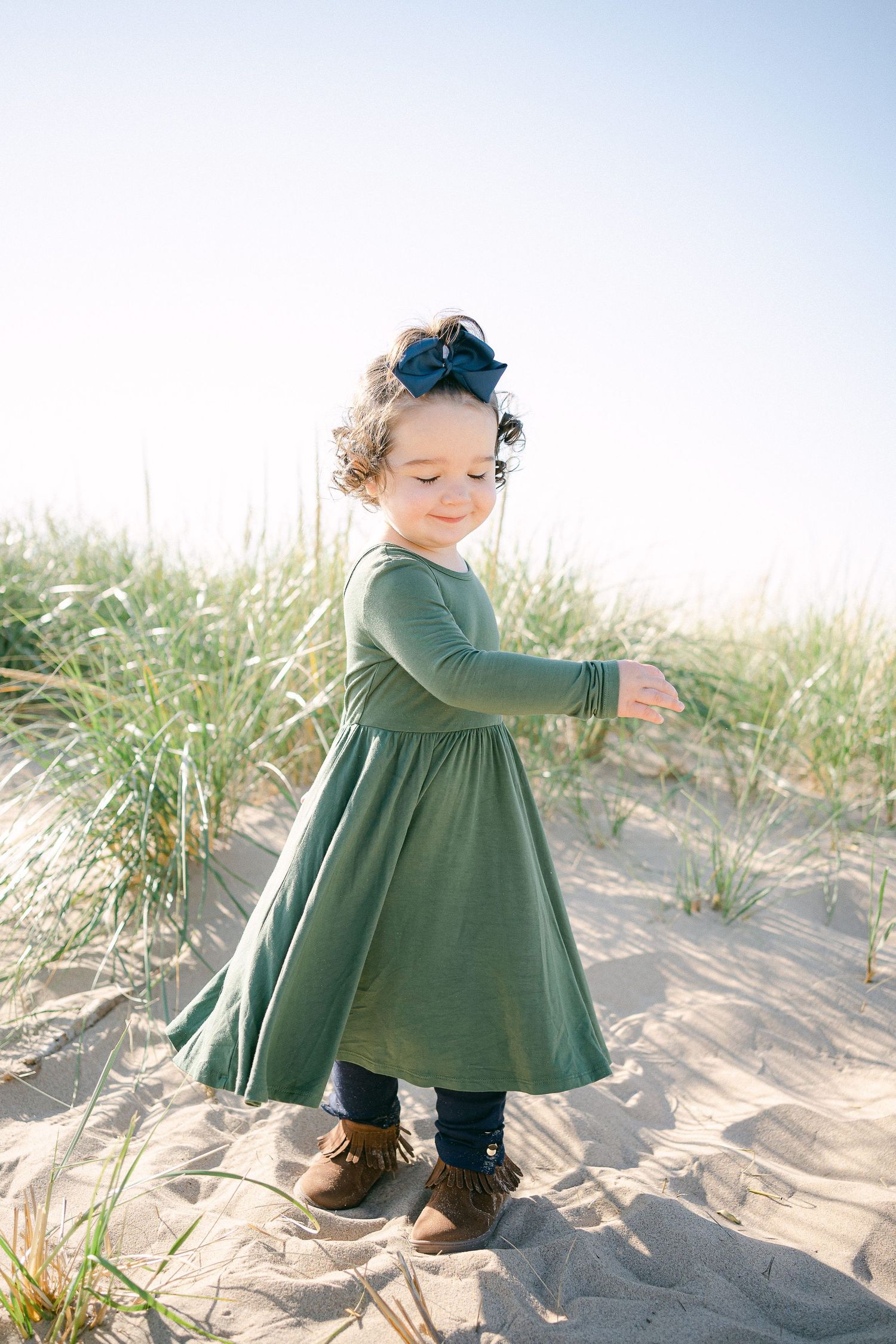 A small figure in a green dress plays in sandy beach dunes with tall grass during golden hour.