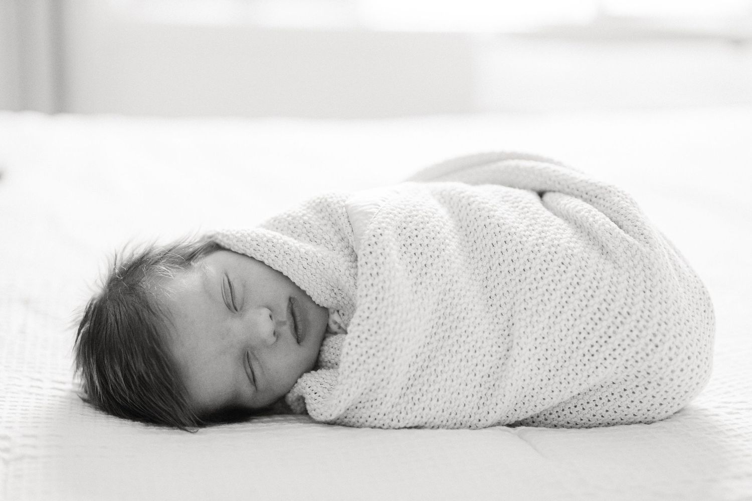 A series of four black and white photos showing a newborn baby swaddled in white blankets laying on a soft surface.