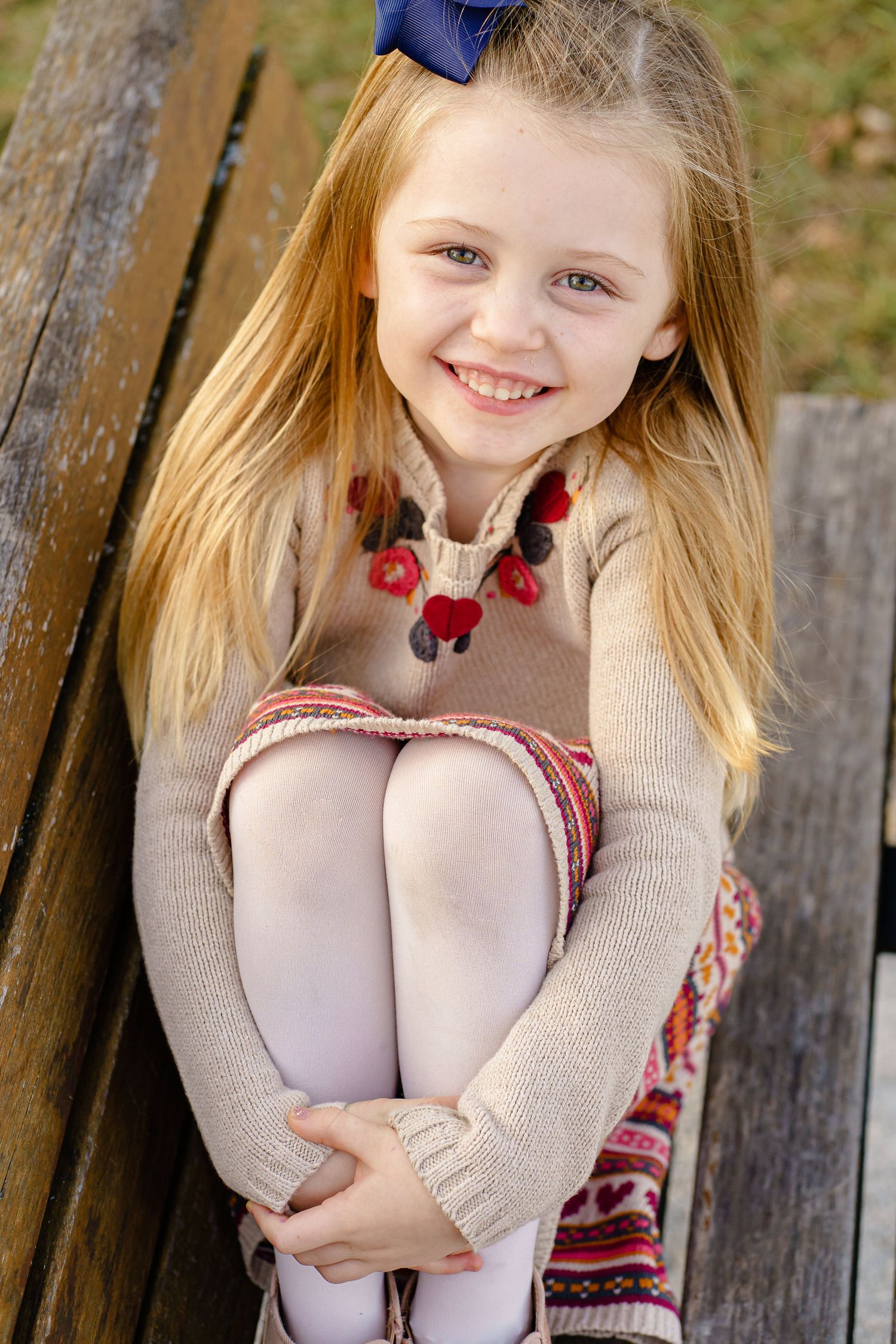 A young child with blonde hair wearing a beige sweater and red necklace sits on a wooden bench outdoors.