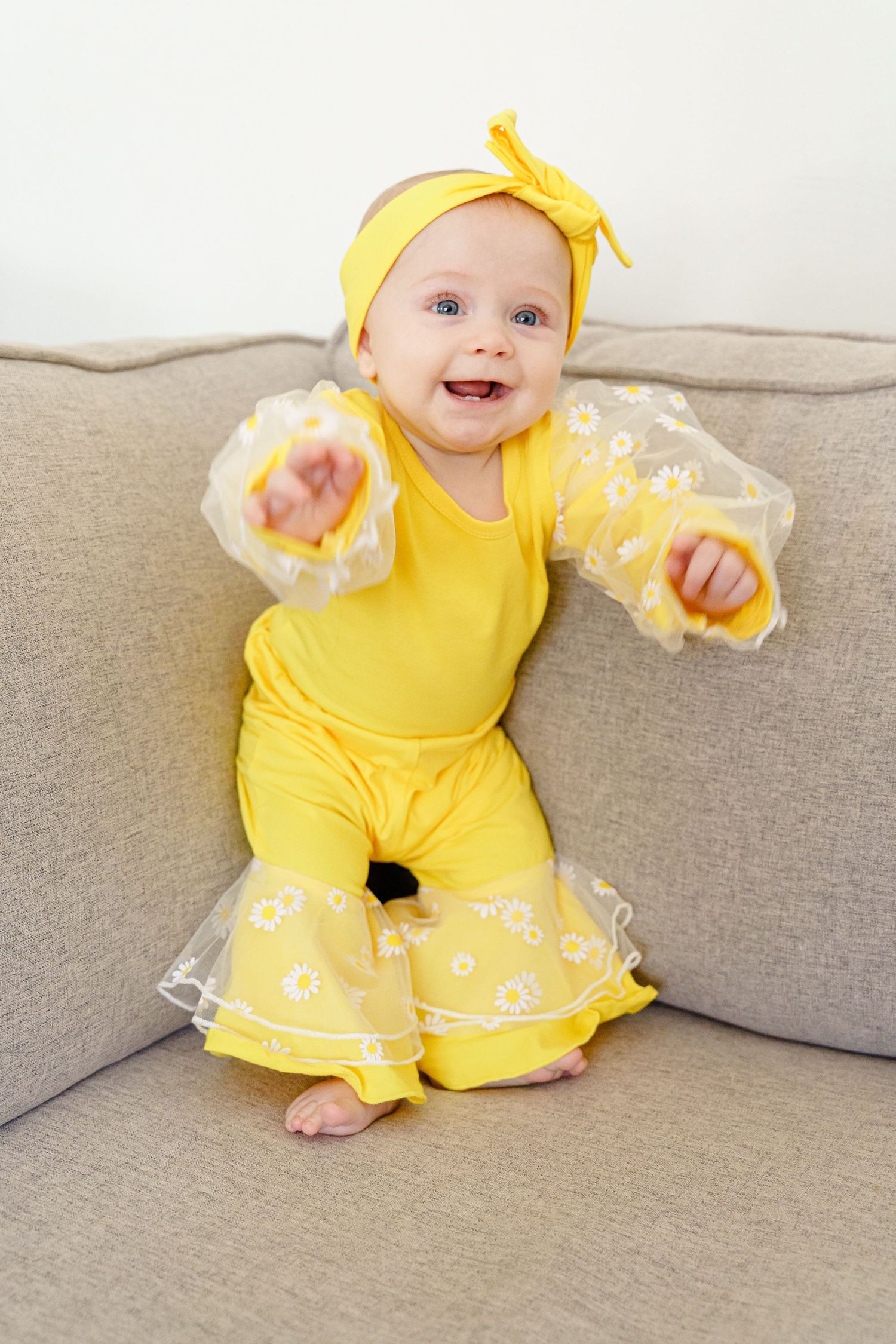 A small child wearing a bright yellow dress with white trim sits happily on a beige couch.