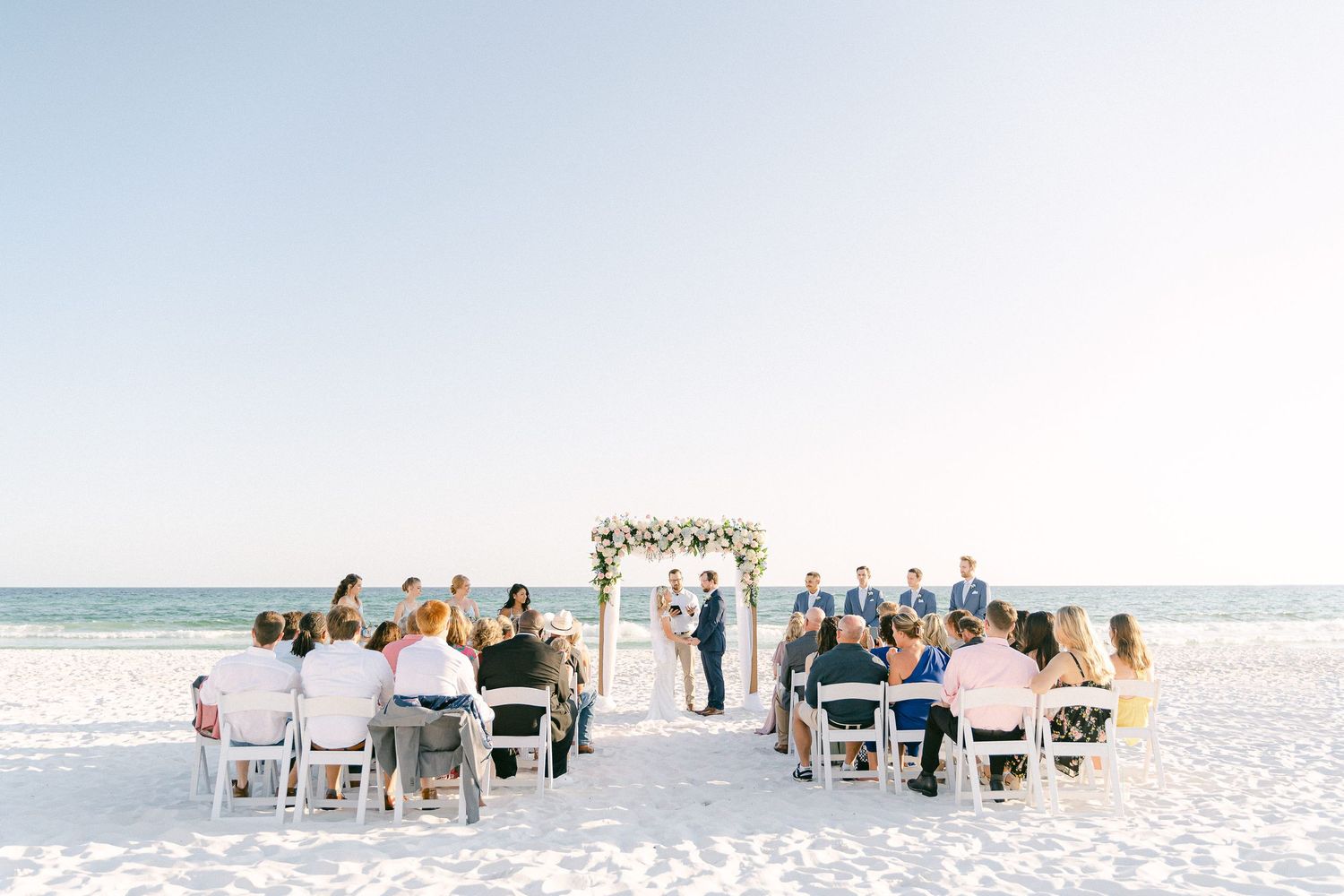 Romantic beach wedding ceremony with guests seated on white chairs facing couple at floral arch, ocean backdrop, clear sky in Destin, FL