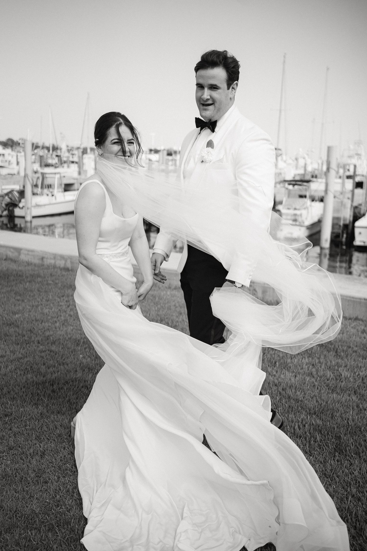 Black and white photo of a bride and groom laughing near a marina, her wedding dress flowing in the wind in Miami, FL