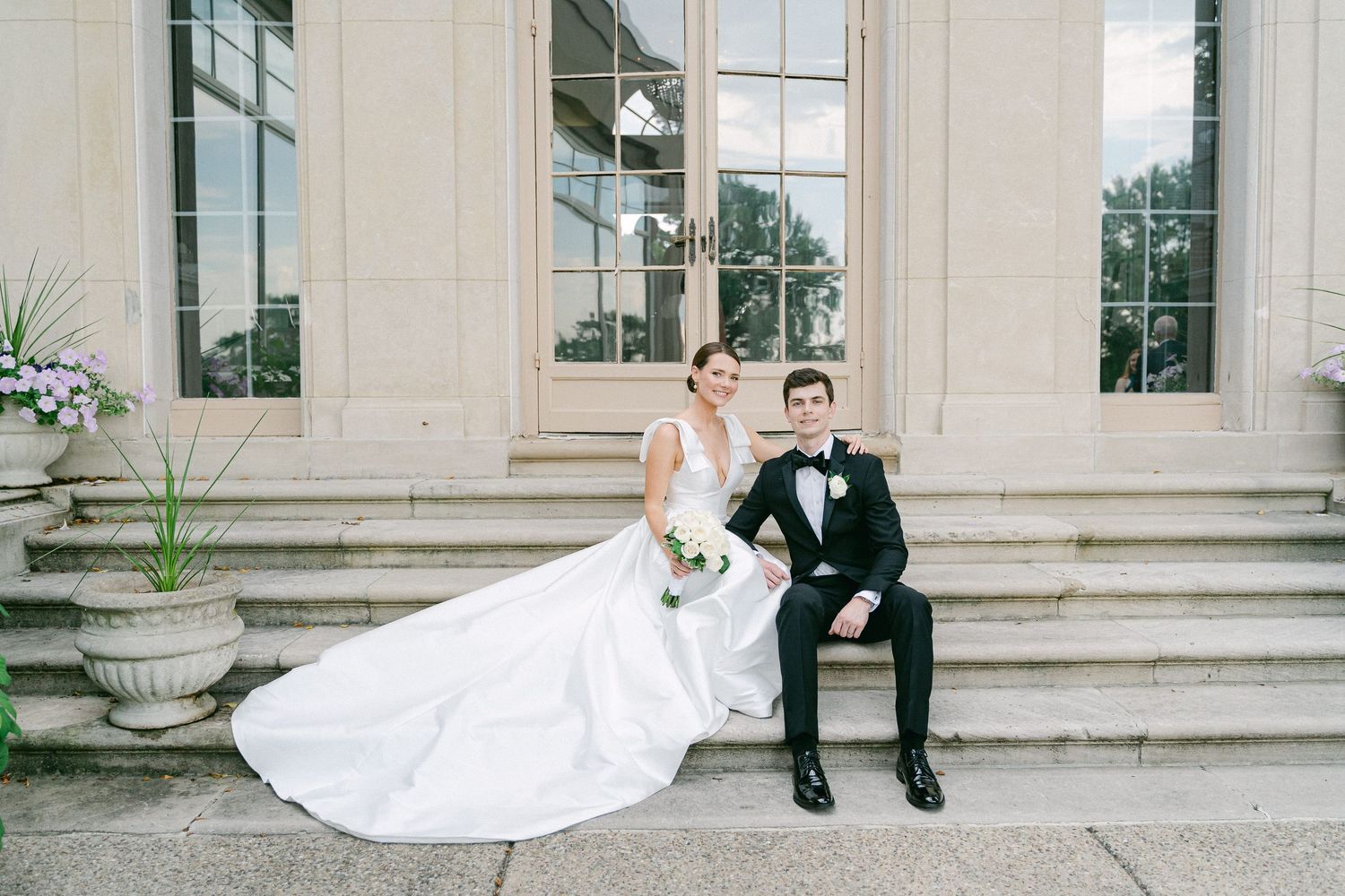 Bride and groom sitting on stone steps of elegant venue, bride in flowing white gown, groom in classic black tuxedo in Philadelphia, PA