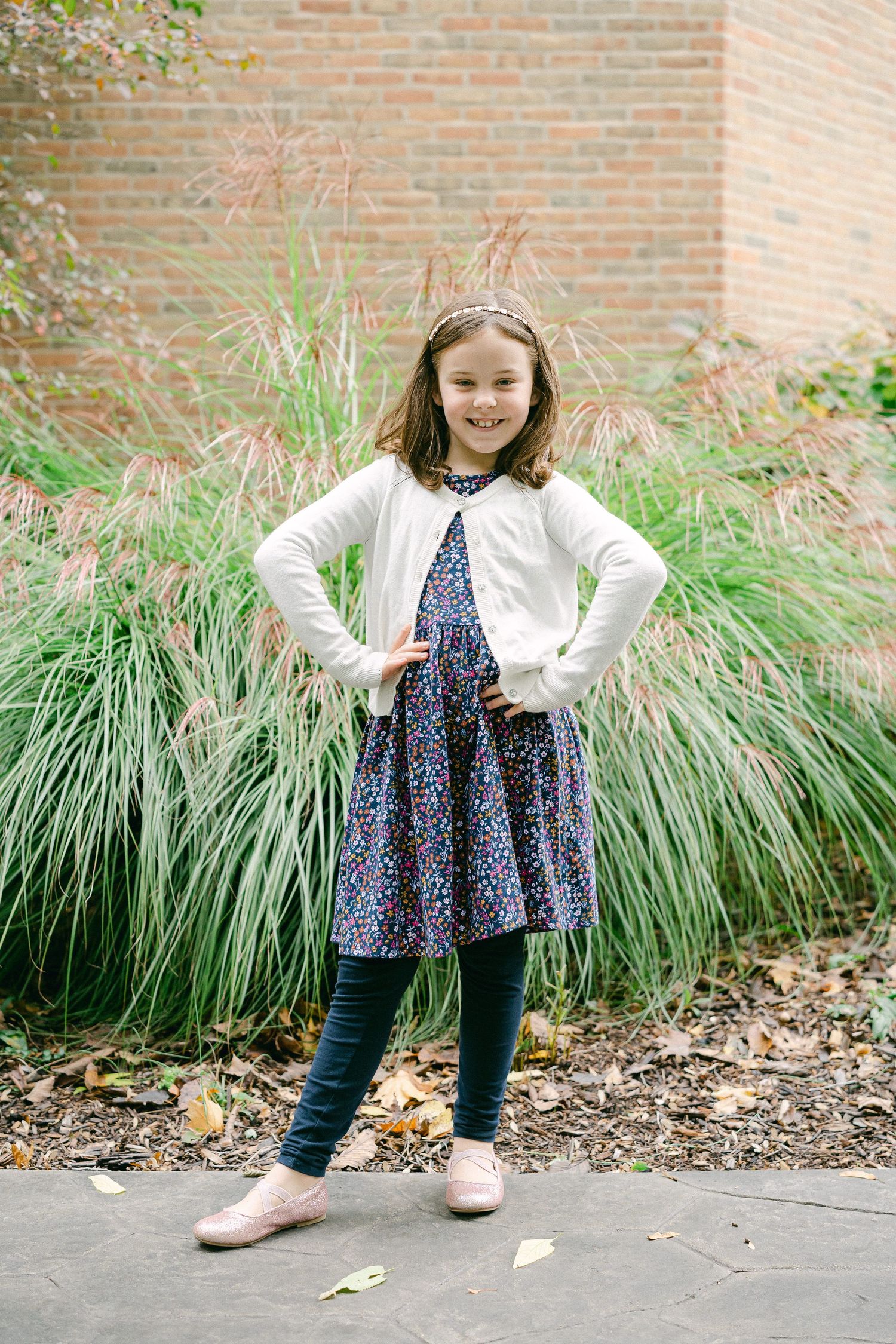 Child wearing a floral dress and white cardigan poses against a brick wall with ornamental grasses.