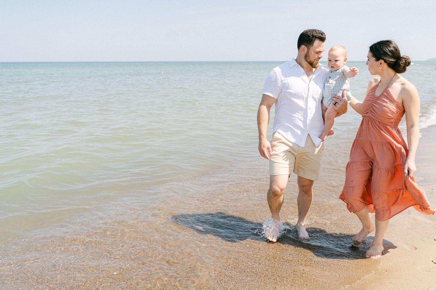 A couple in white and coral outfits walks hand in hand along a sandy beach by the ocean.