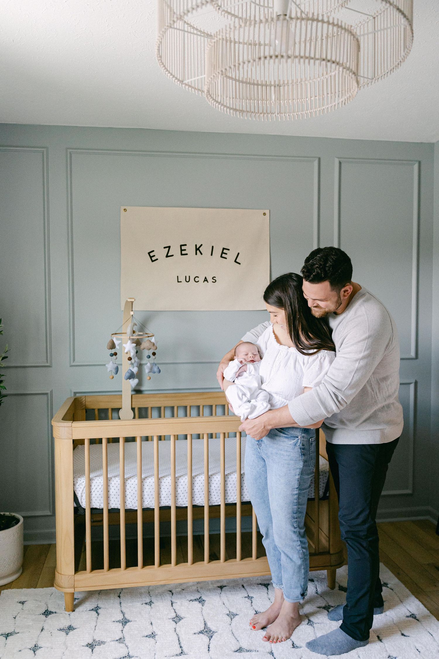 New parents hold their baby in a nursery with sage green walls and wooden crib displaying name art.