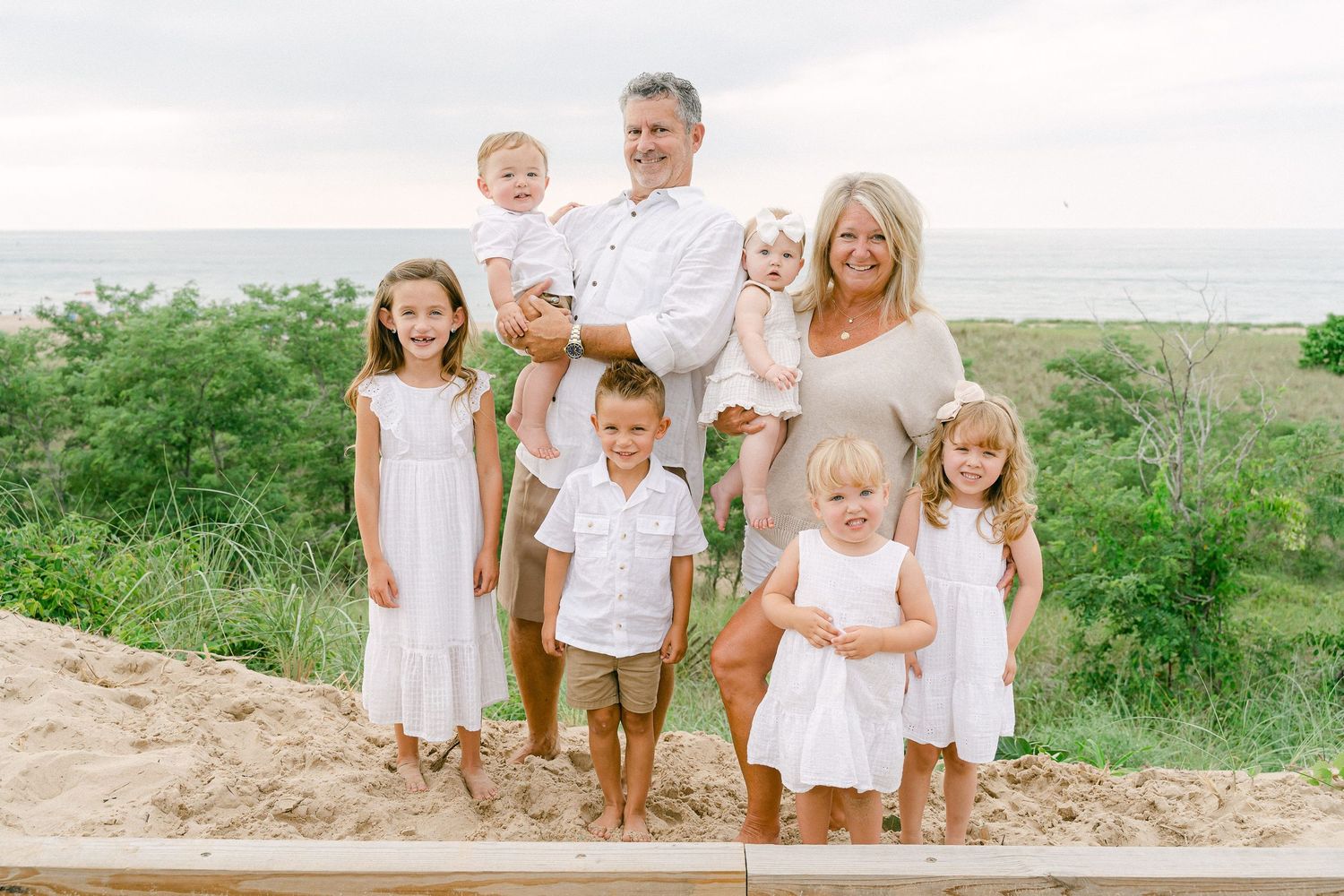 Extended family dressed in white and beige clothing gathers for outdoor portrait session near beach.