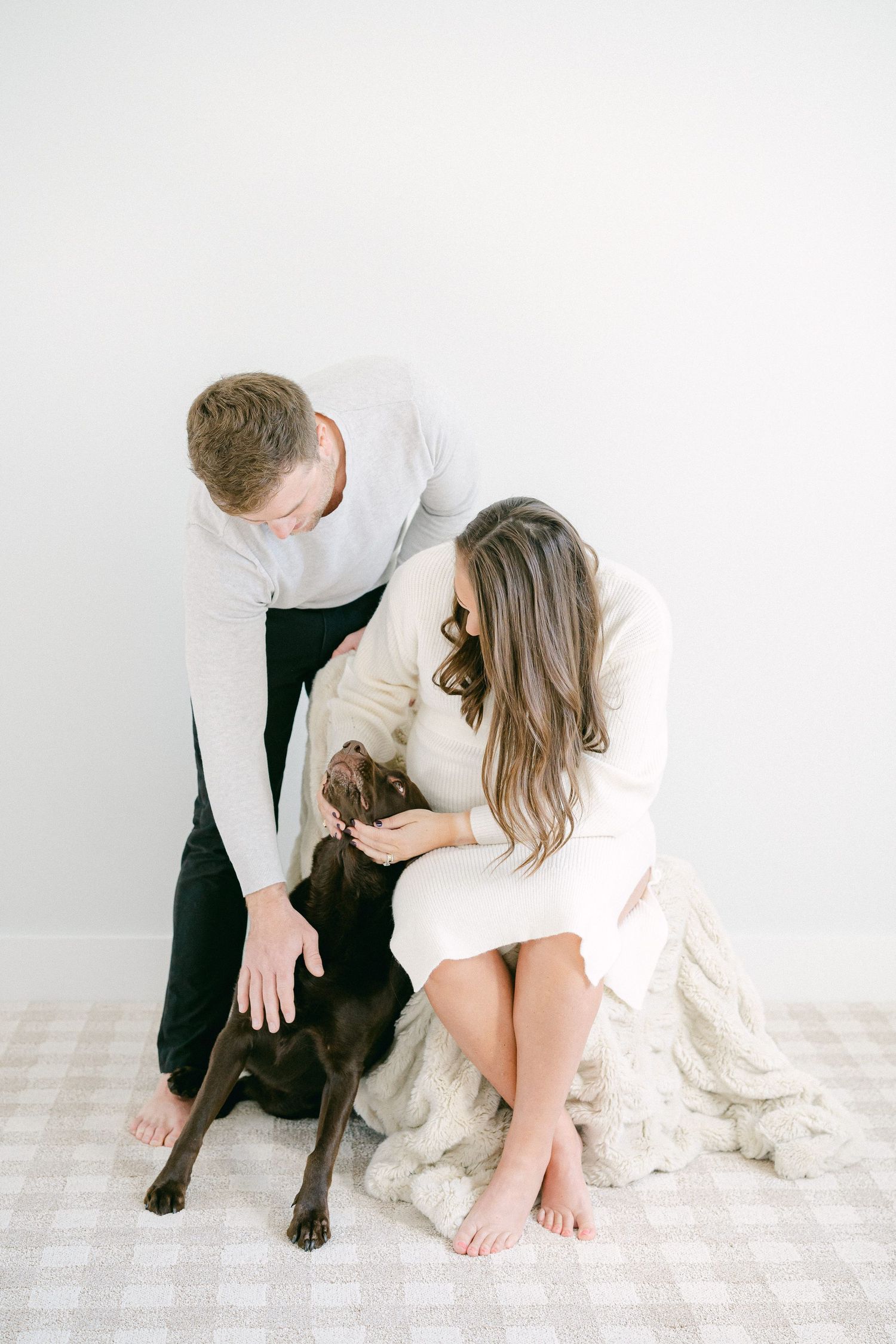 A couple in white sits on the floor with their chocolate labrador retriever in a bright studio setting.
