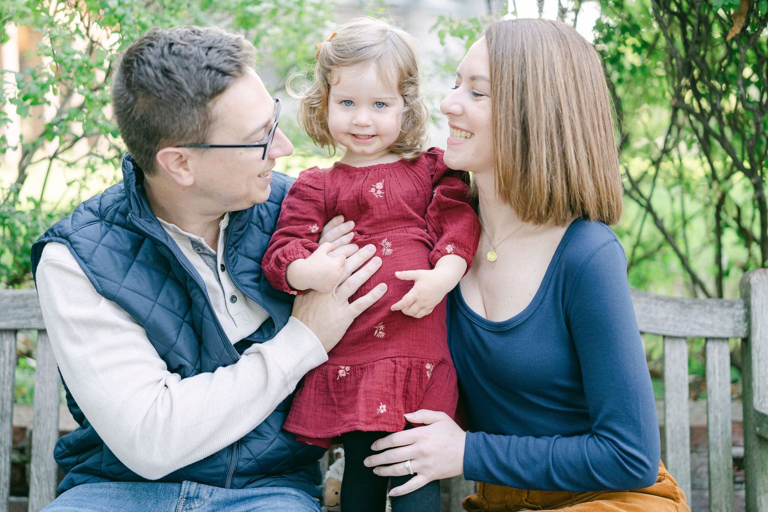 Family wearing coordinating navy and burgundy outfits sits together on wooden bench during autumn outdoor photo session.