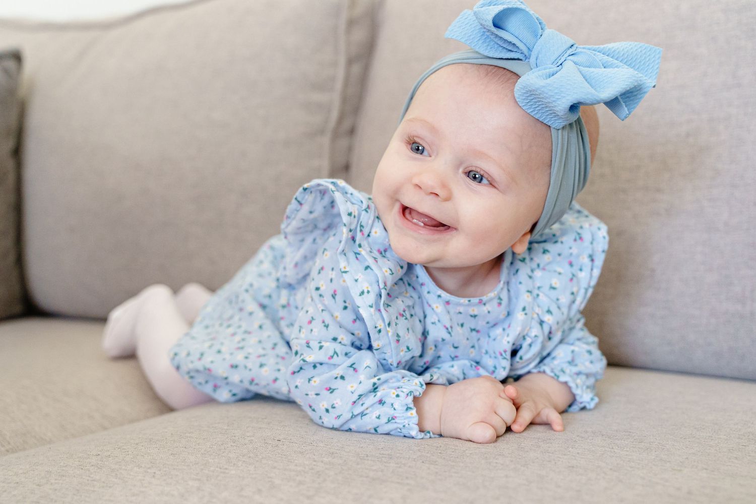 A baby wearing a light blue floral outfit and matching bow lies smiling on a gray couch.