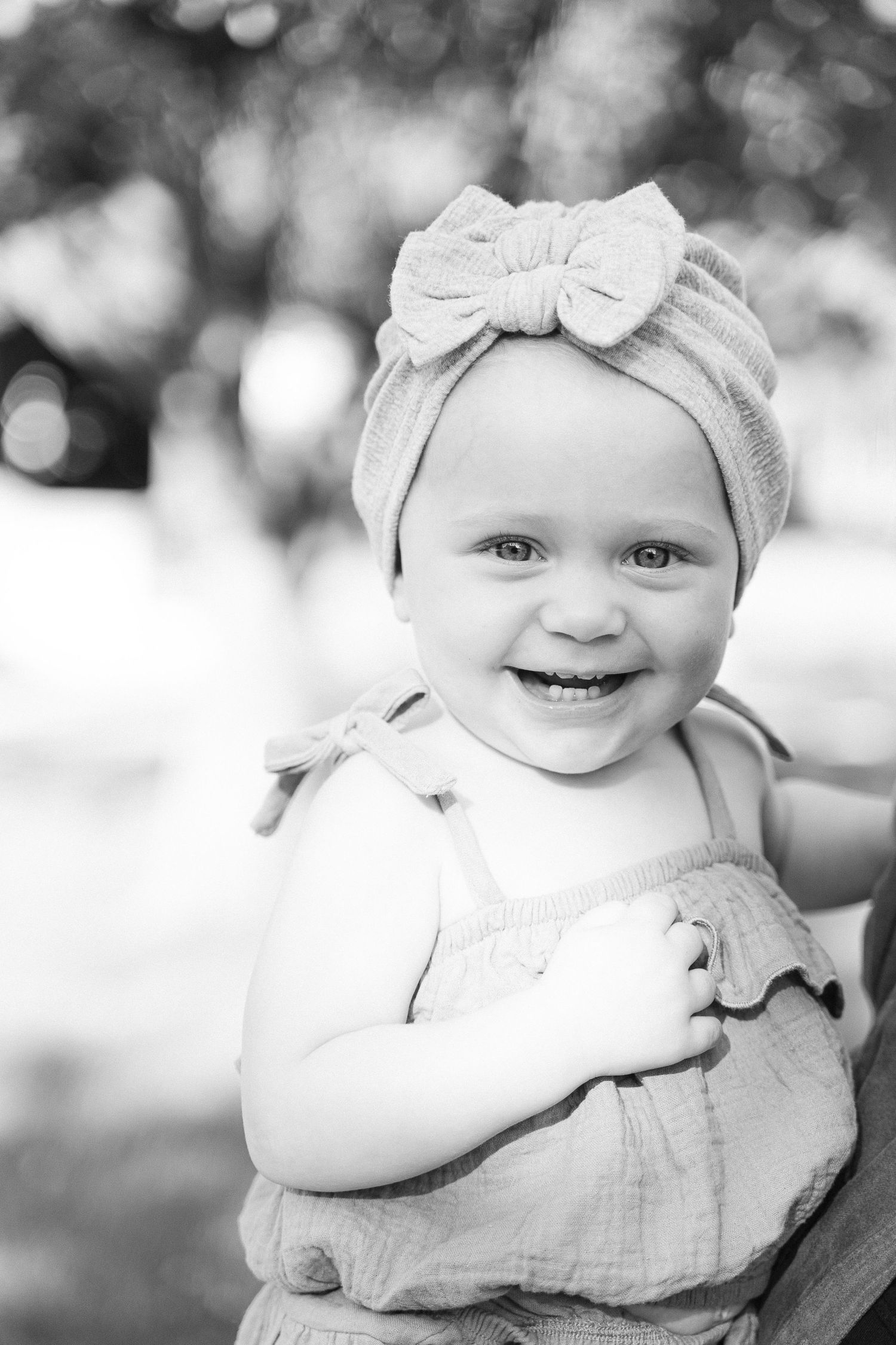 Black and white portrait of a smiling baby wearing a bow headband and linen romper outdoors.