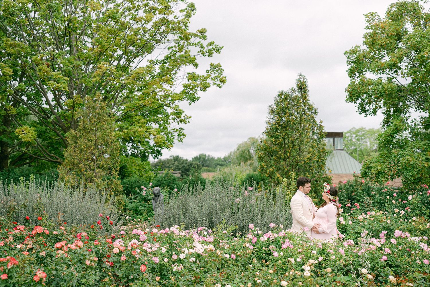 A panoramic view of a lush garden landscape with blooming flowers and trees under a cloudy summer sky.