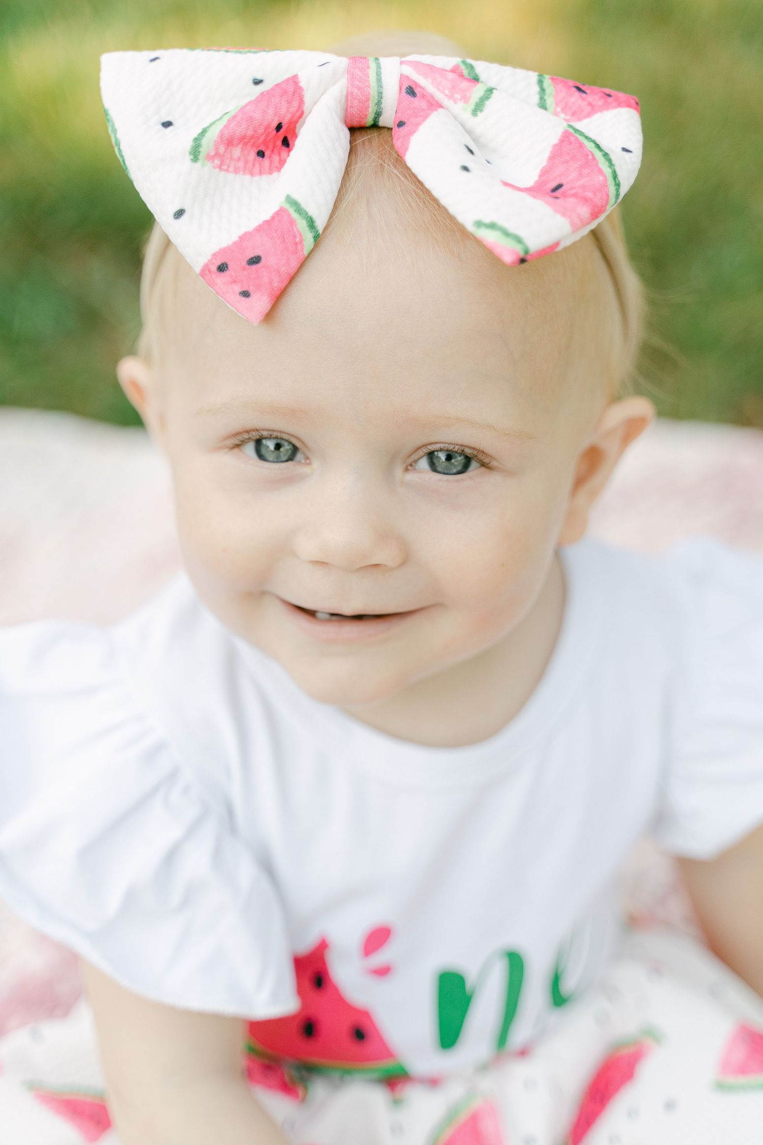 A sweet summer-themed outfit with a watermelon bow and white shirt against a green outdoor background.