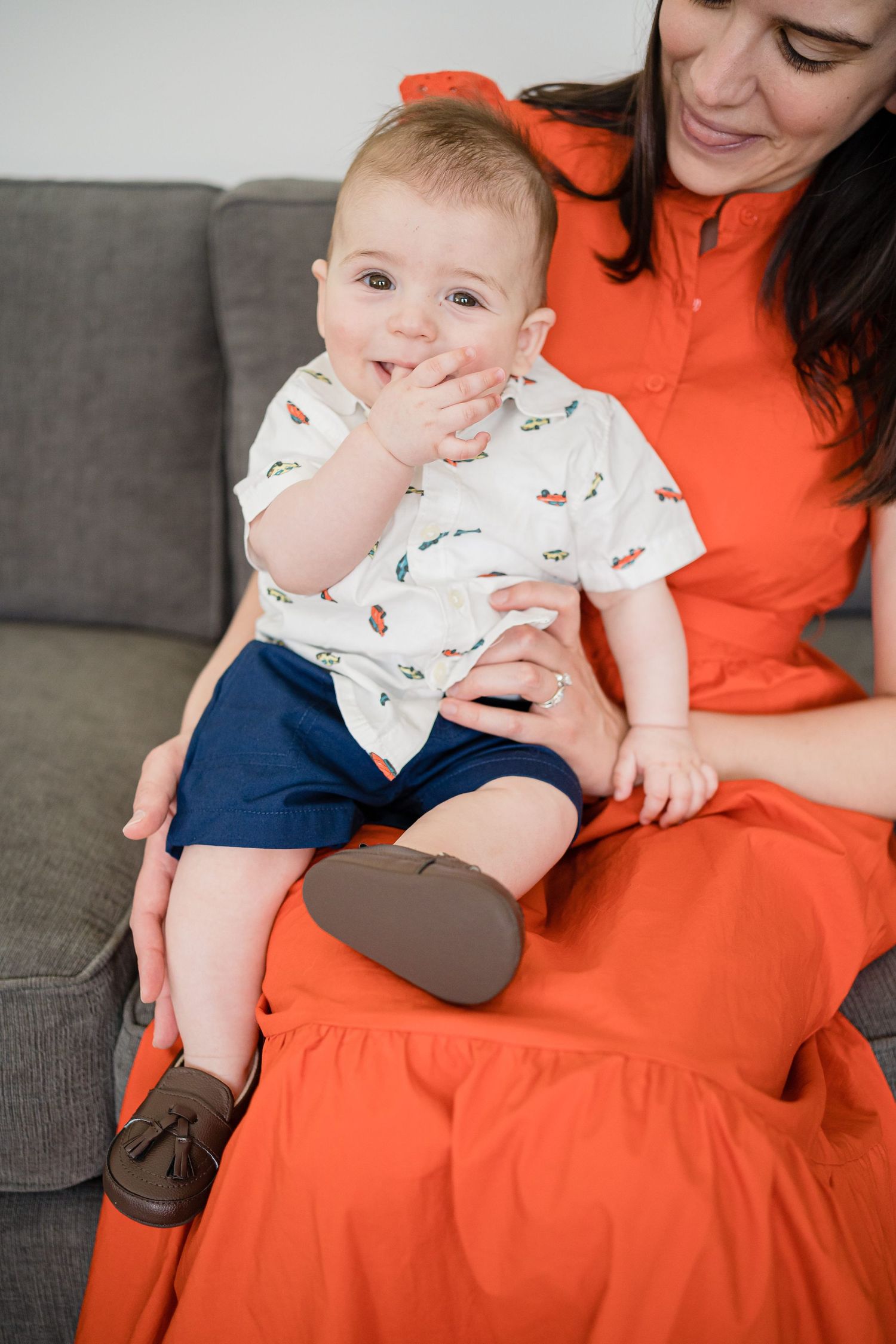 Baby wearing a patterned onesie sits on orange fabric while being supported by an adult.