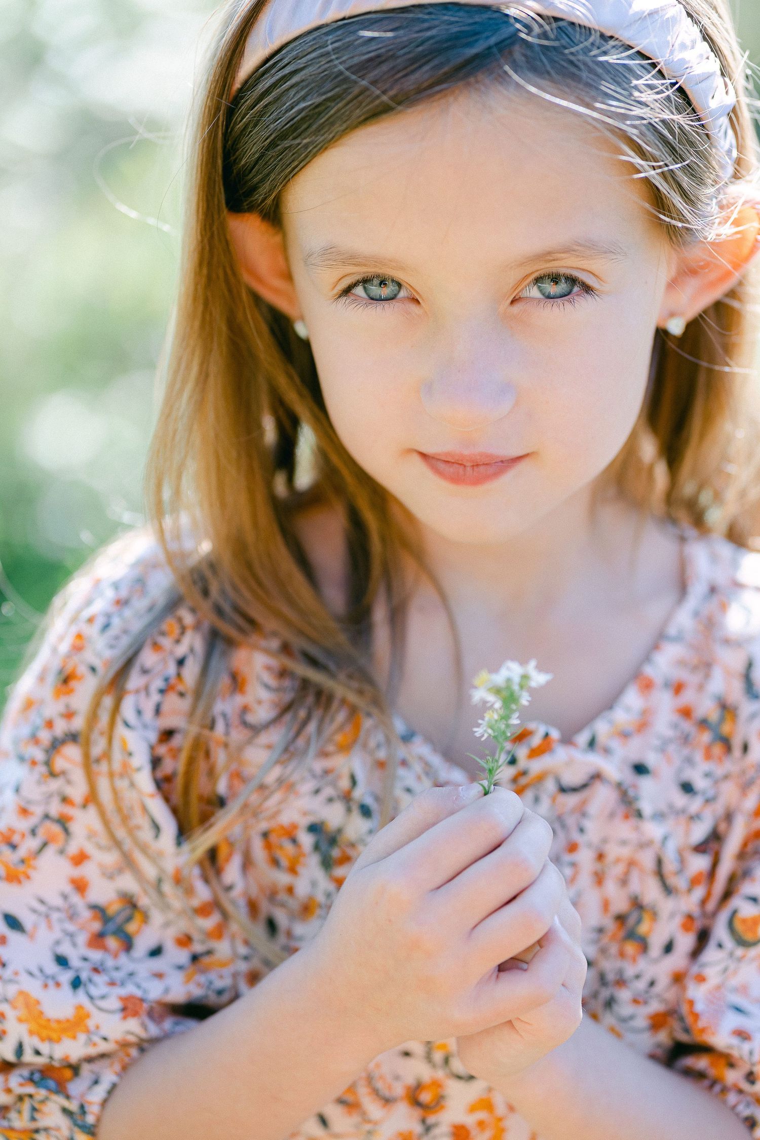 Young child in floral dress holding small white wildflower against soft bokeh background in sunlight.