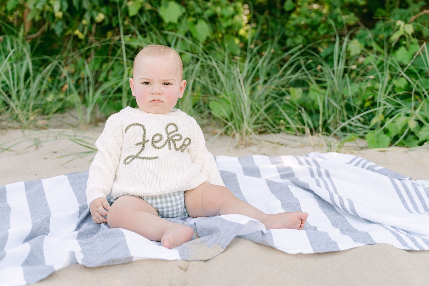 Baby sitting on striped blanket outdoors wearing cream sweater and plaid shorts in garden setting.