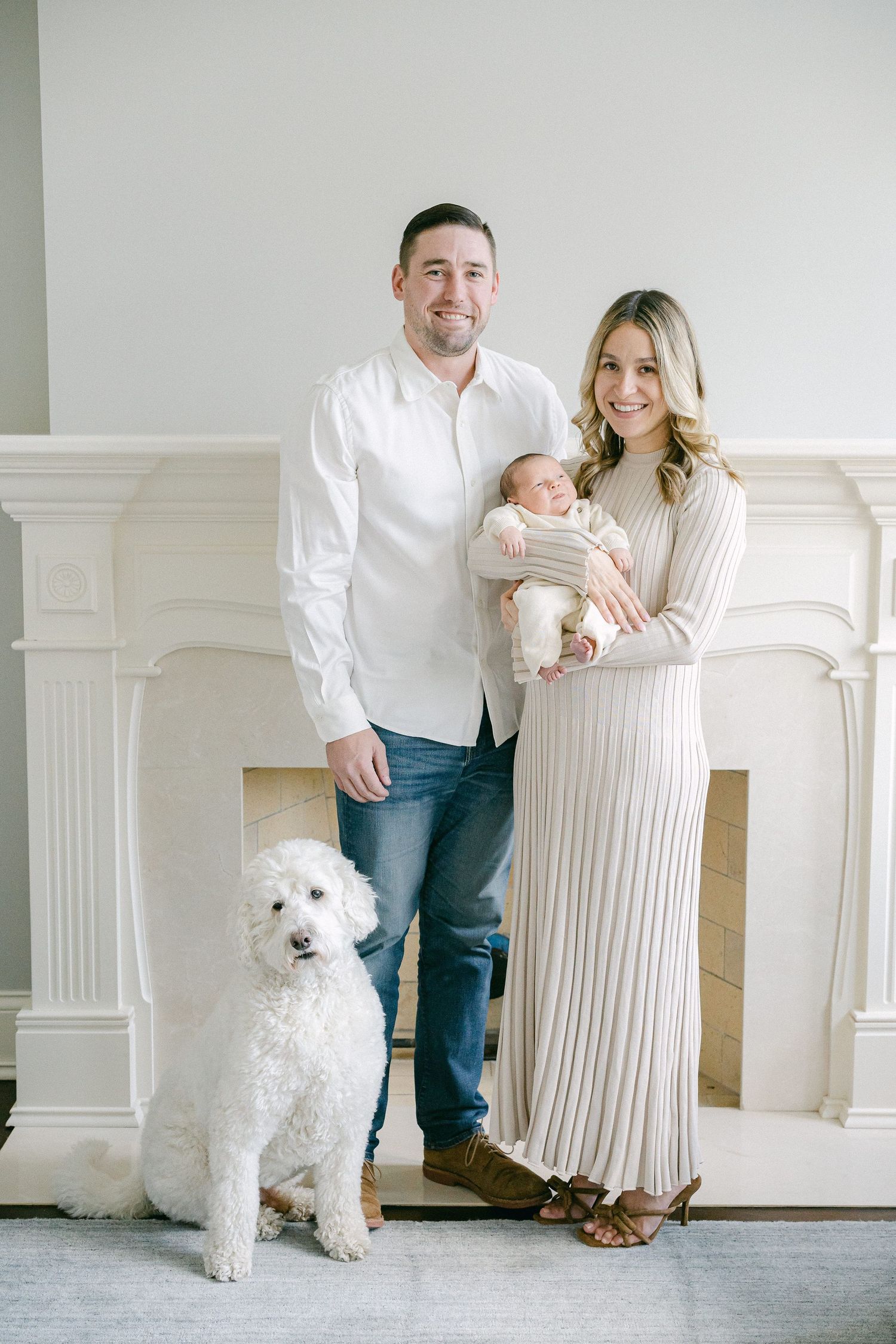 Family portrait in front of white fireplace with fluffy white dog sitting beside them in coordinated neutral outfits.