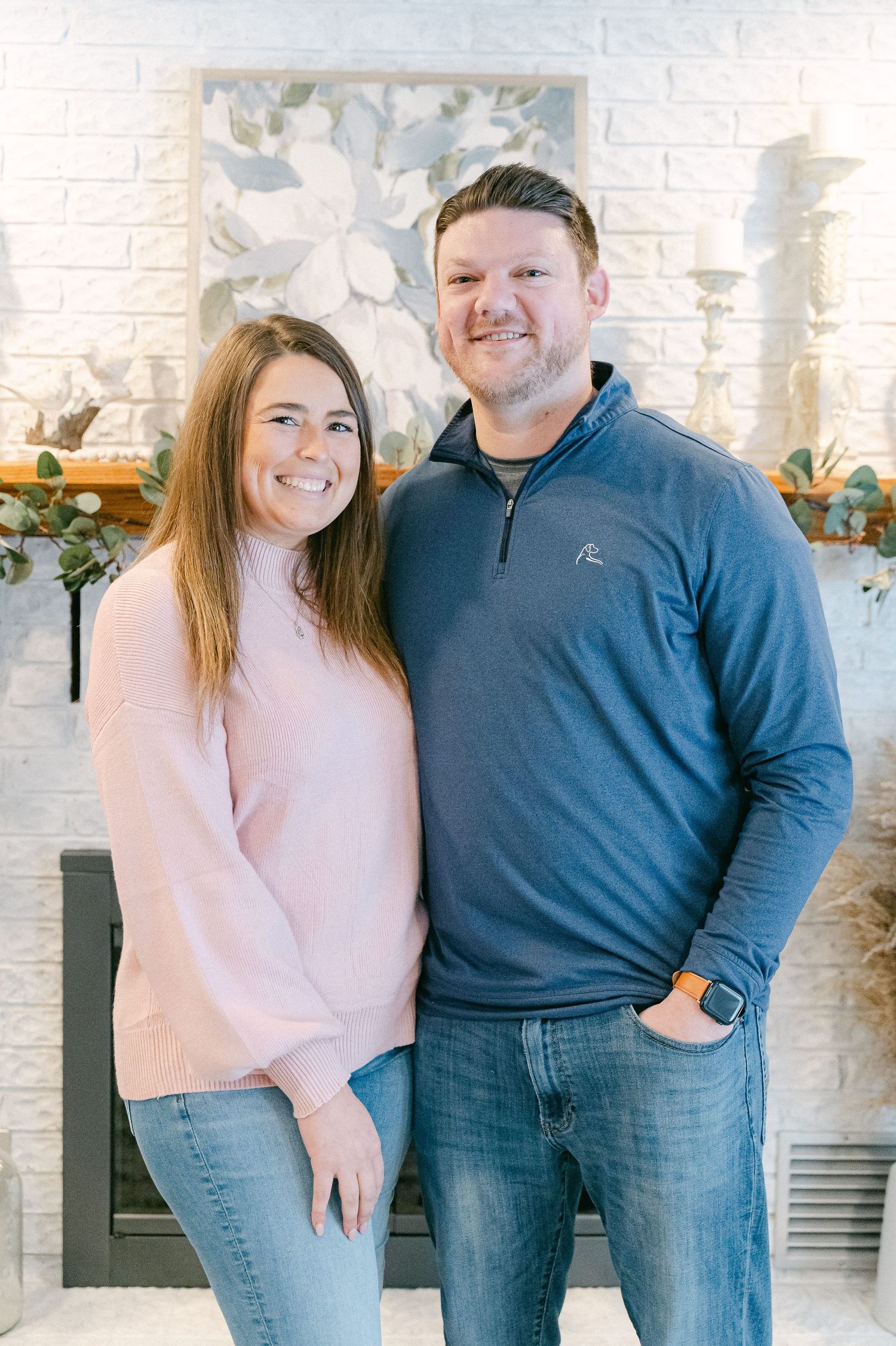 Couple wearing matching pink and blue sweaters pose together against white brick wall in casual indoor photoshoot.