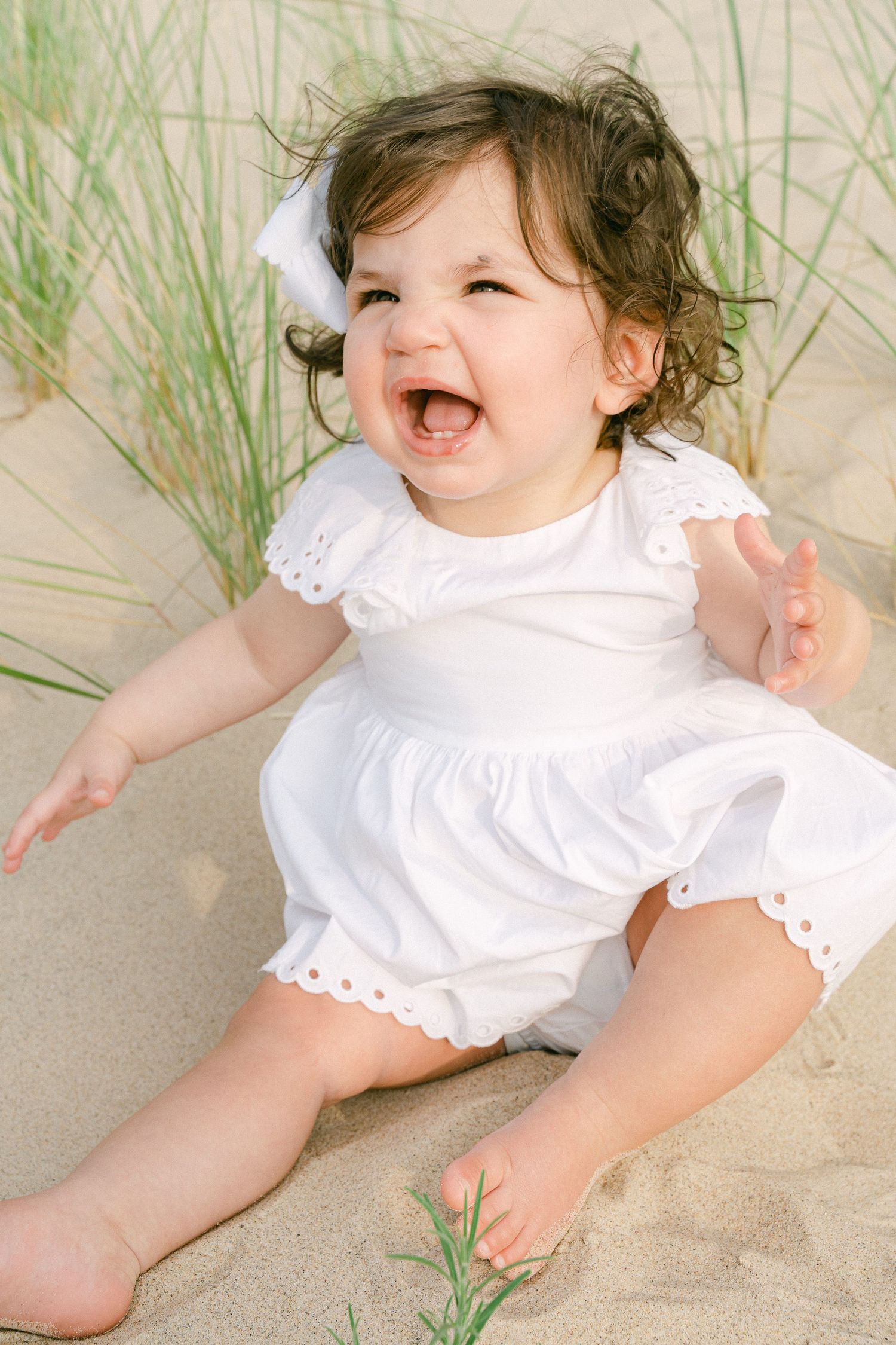 Baby in white eyelet dress laughing while sitting on sandy beach with sea grass.