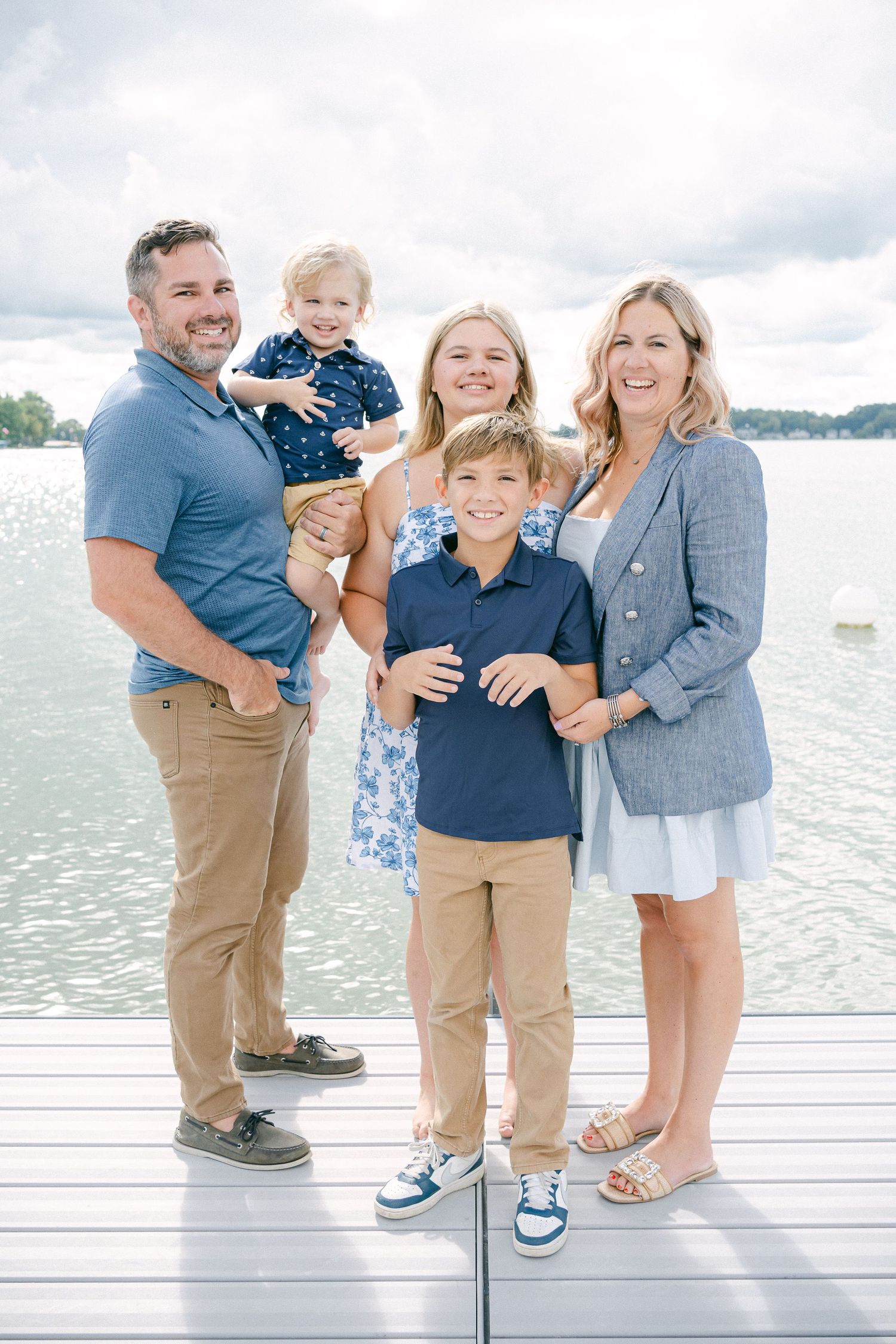 A family poses together on a white boardwalk by the water wearing coordinated blue and neutral outfits.