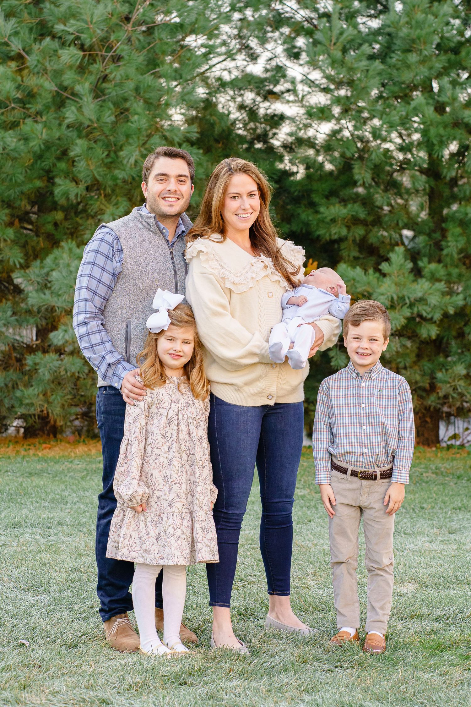 A family of five poses together outdoors in coordinated casual outfits against green foliage in a backyard setting.