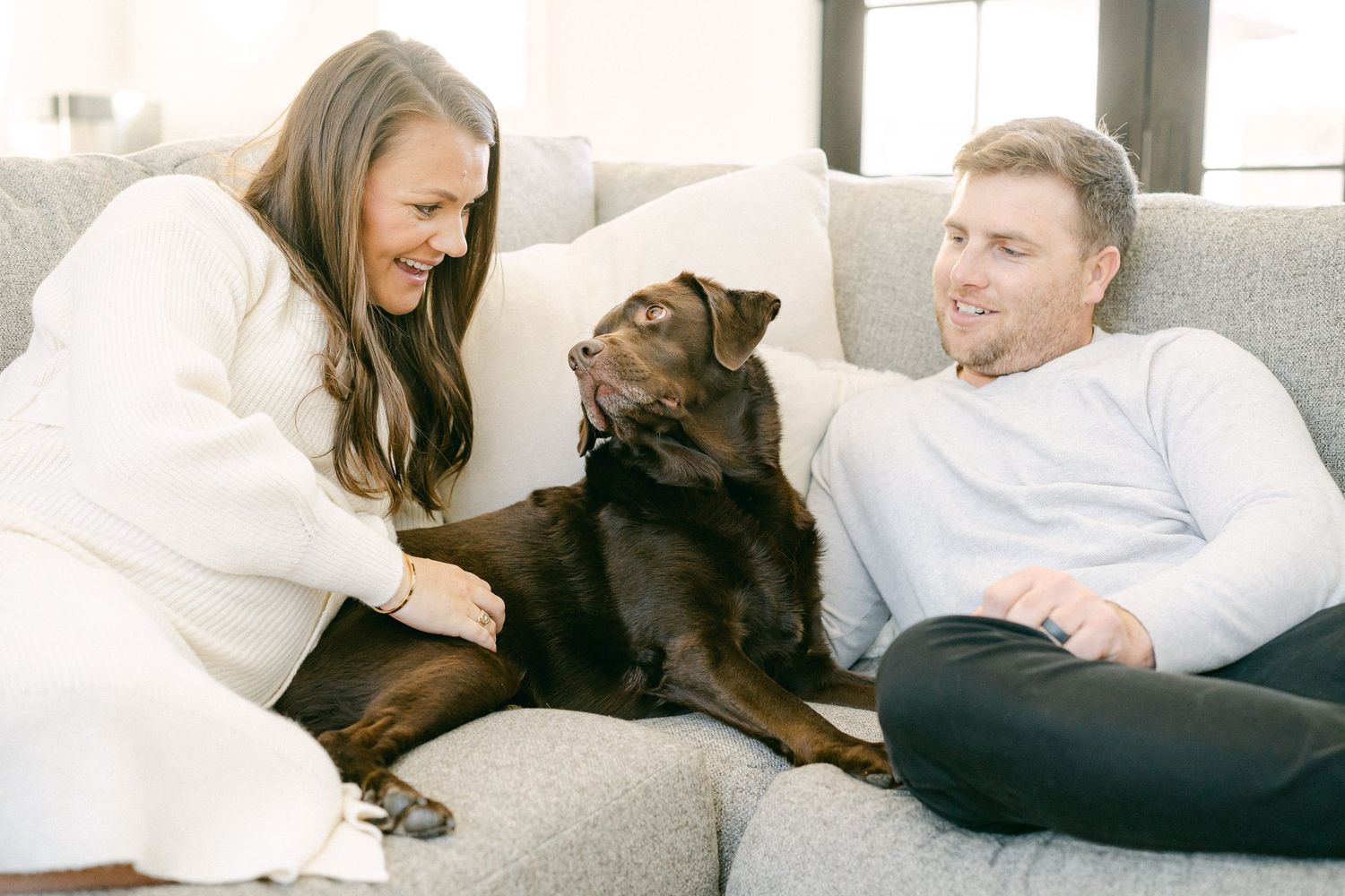 Happy couple relaxes on grey couch with their chocolate lab.