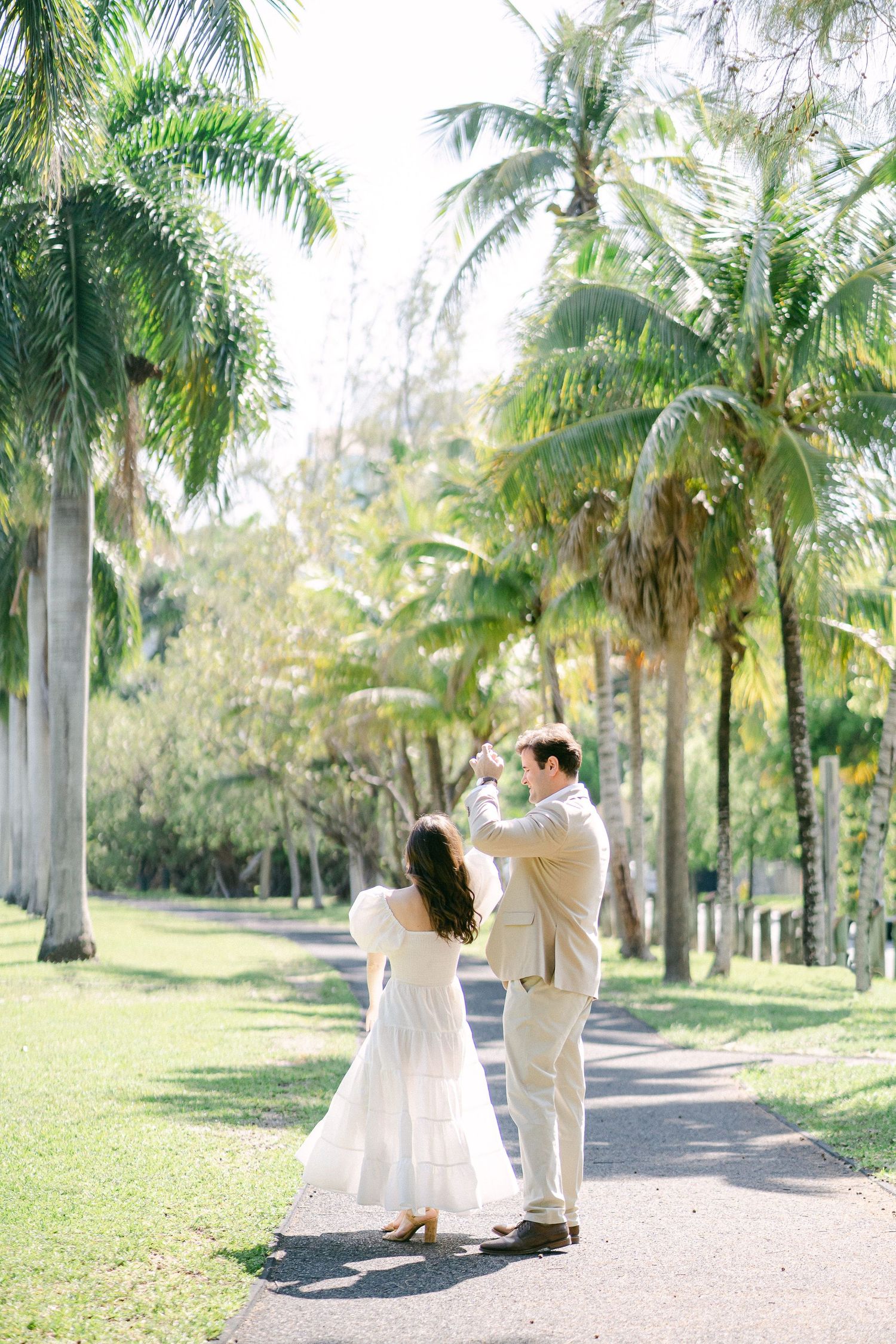 Couple dancing on a palm-lined path, woman in white dress and man in beige suit, tropical garden setting in Miami, FL