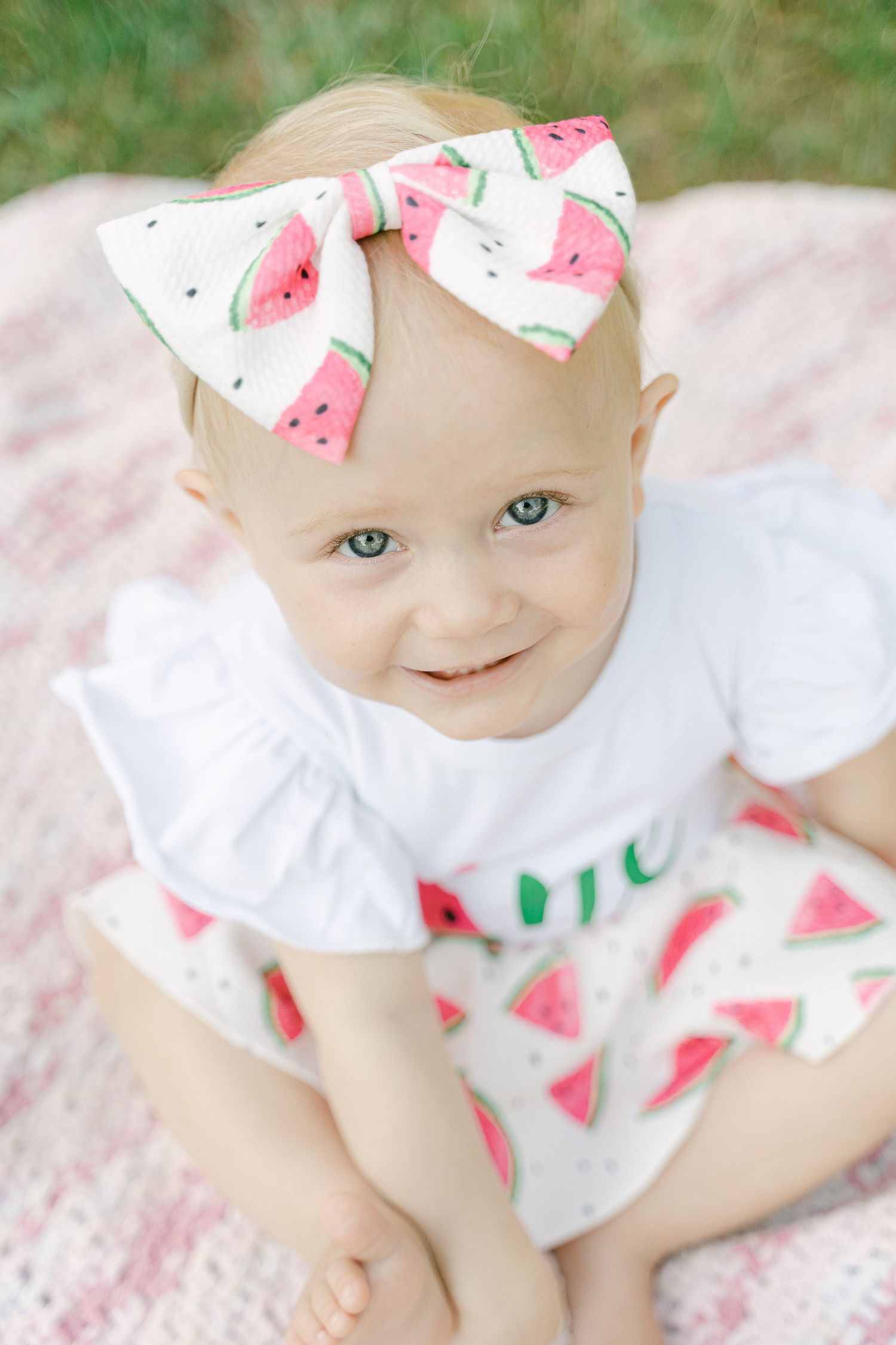 Baby wearing watermelon print bow and white outfit lying on soft pink blanket.