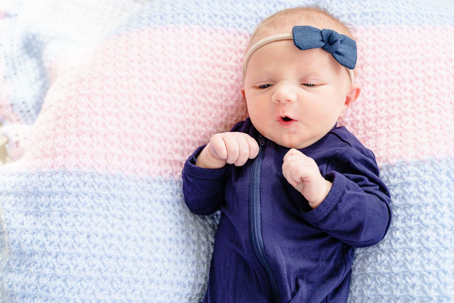 Baby wearing navy blue outfit lies on pink and blue blanket while sucking thumb.