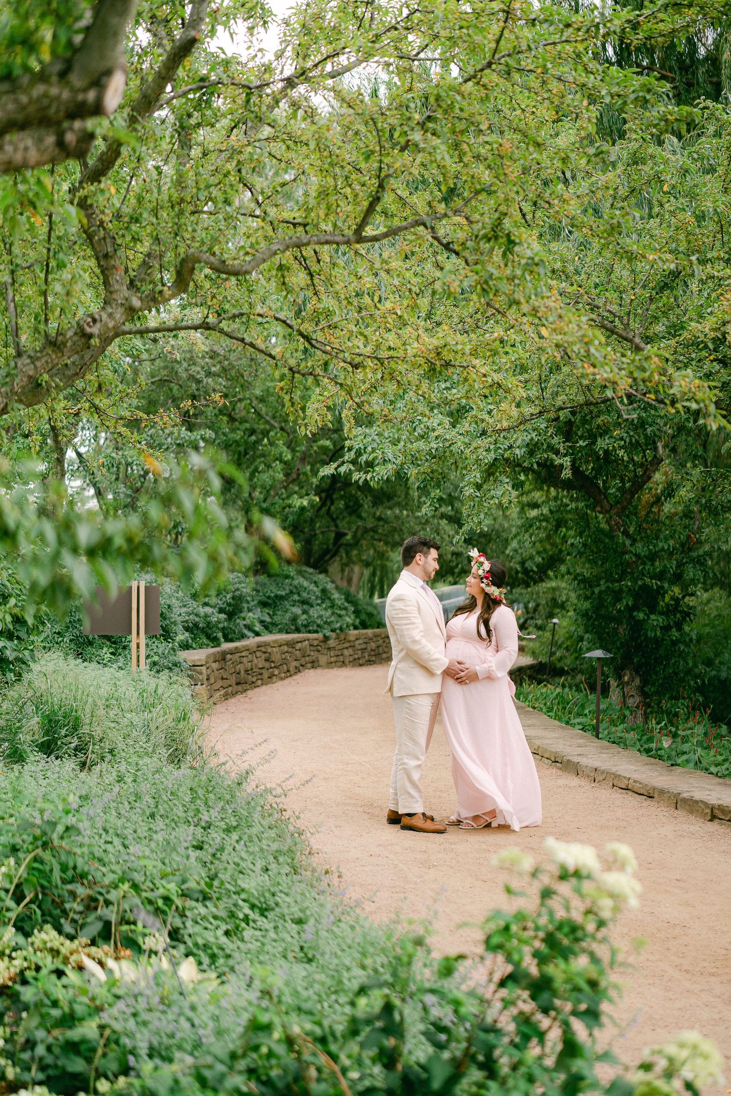 A couple in light colored outfits strolls along a winding garden path surrounded by lush greenery and blooming flowers.