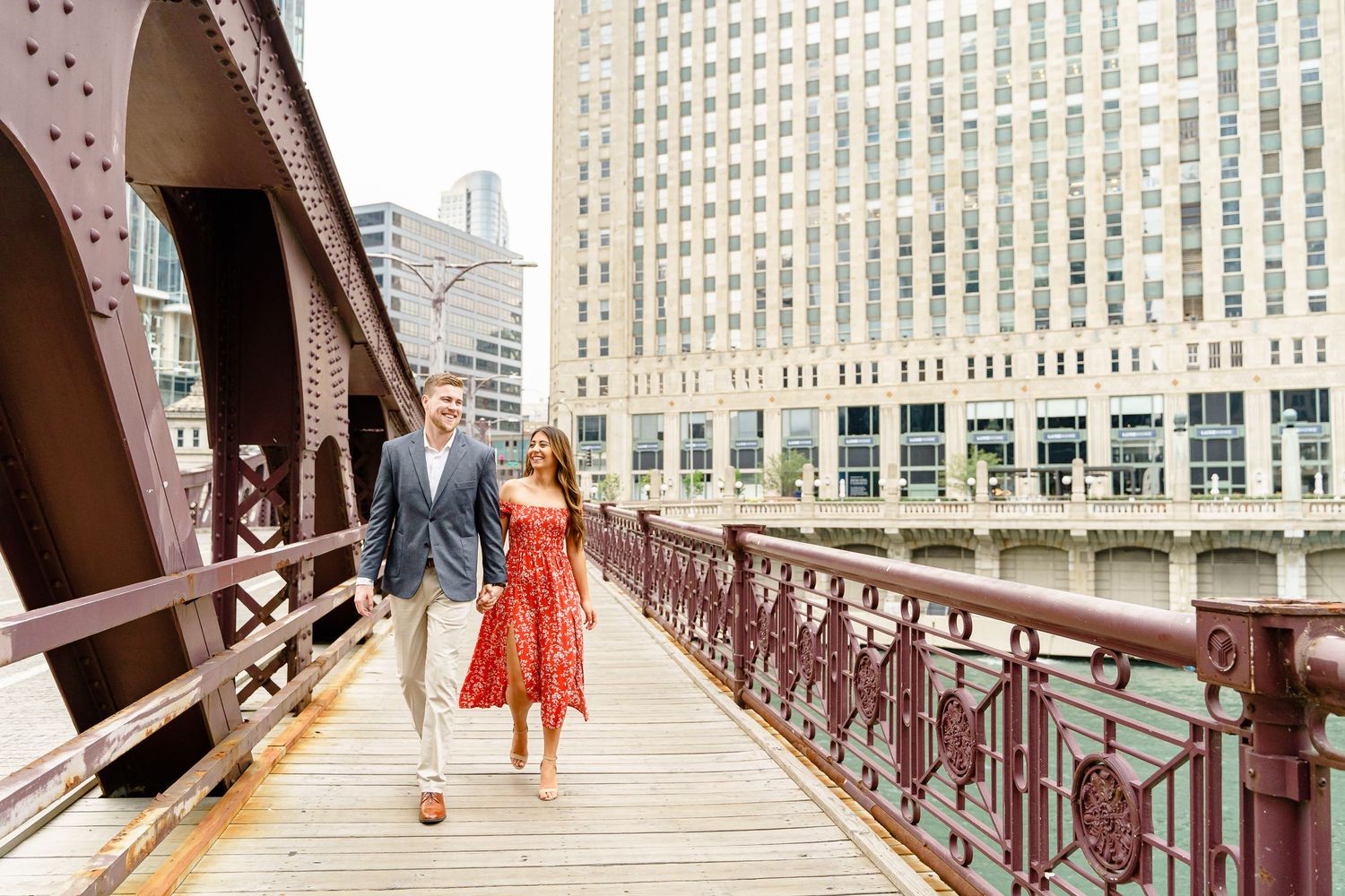 People walk across a vintage iron bridge with ornate railings in an urban setting with tall buildings in the background.