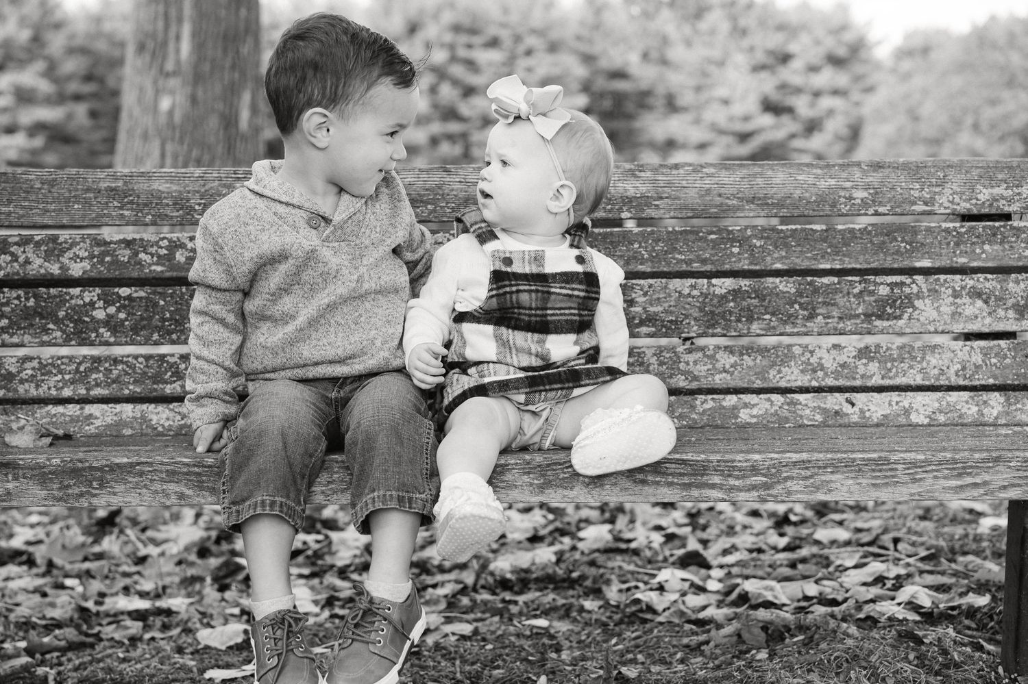 Two children sit on a bench in the park sharing a sweet treat in this nostalgic black and white photograph.