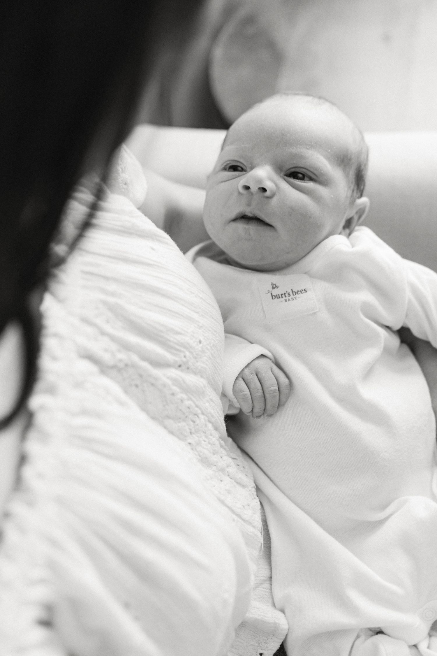 Soft black and white portrait of an infant wearing white clothing while resting on white bedding.