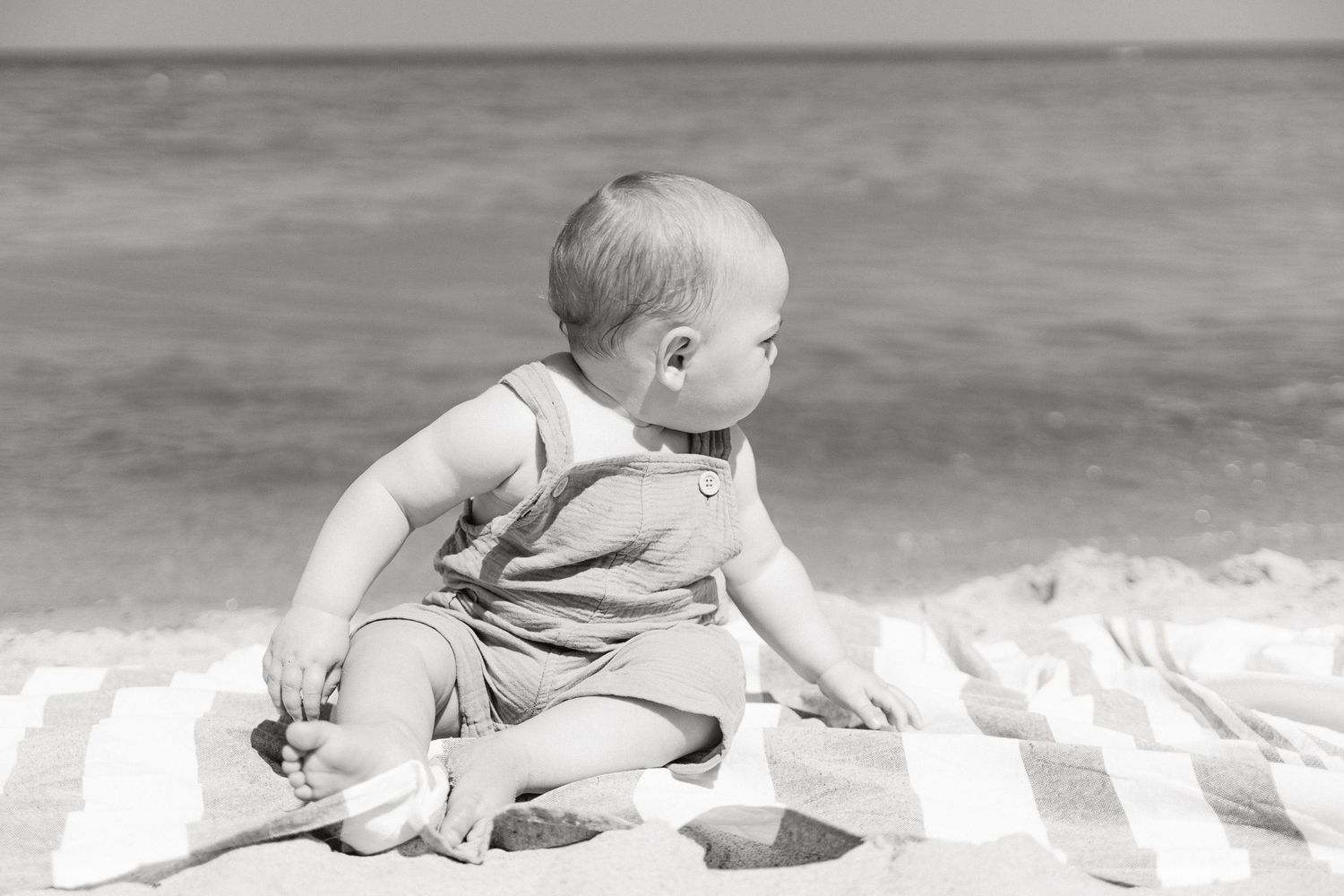 A baby in white clothing crawls along a sandy beach near the ocean waves.