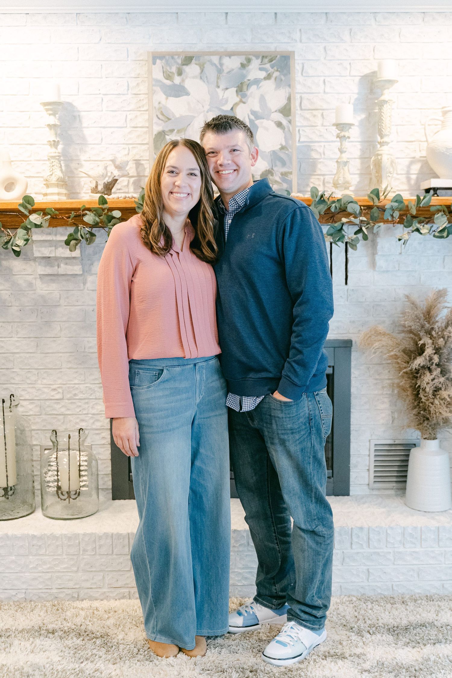 Couple posing in casual outfits against white brick wall with decorative greenery and lanterns.