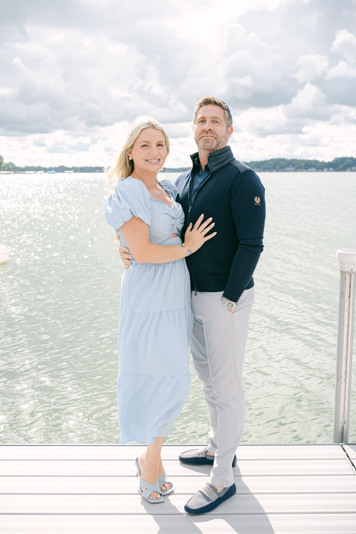 Couple stands together on a white dock overlooking a serene lake on a cloudy day.