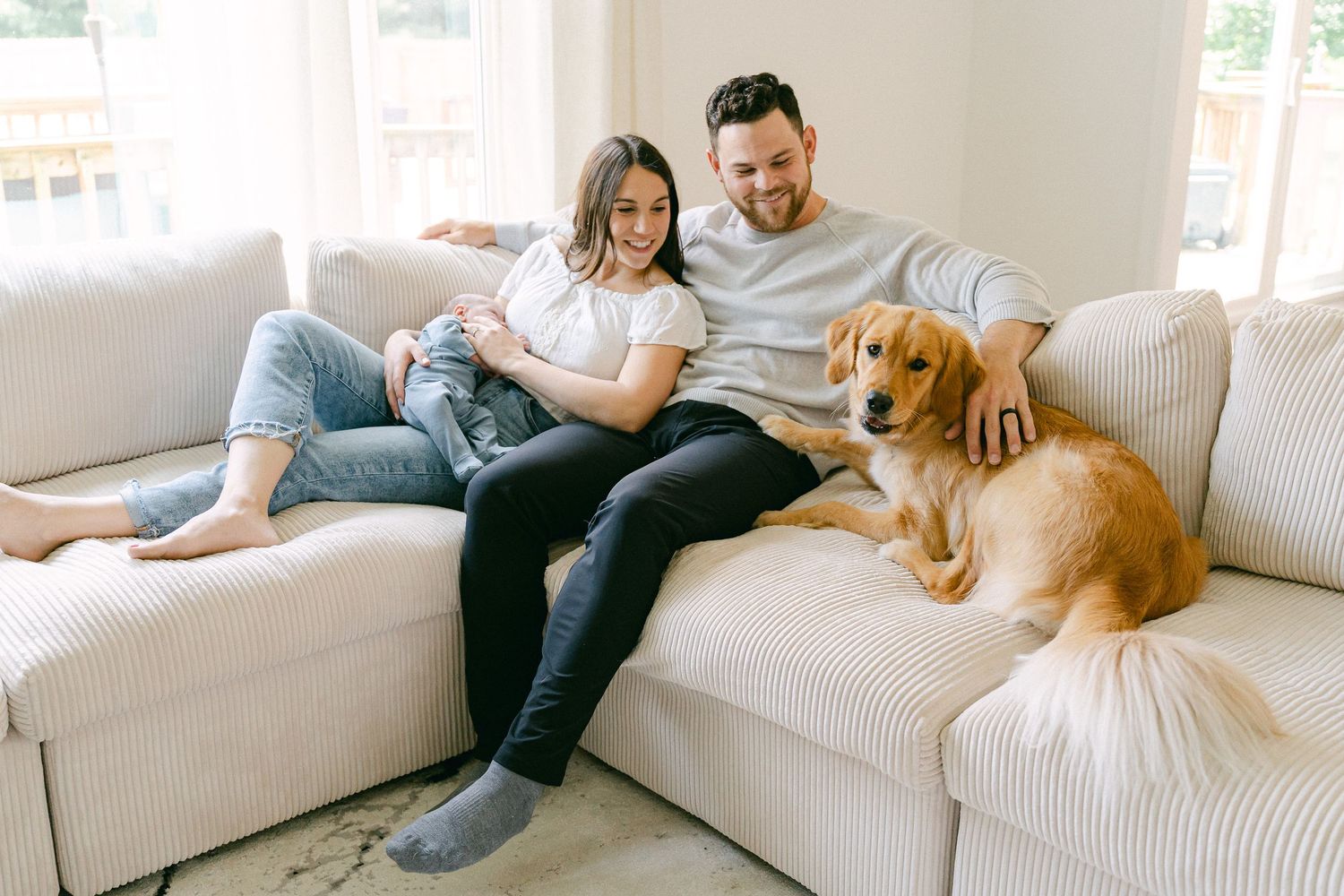 A couple relaxes on a white couch with their golden retriever dog in a bright, modern living room.