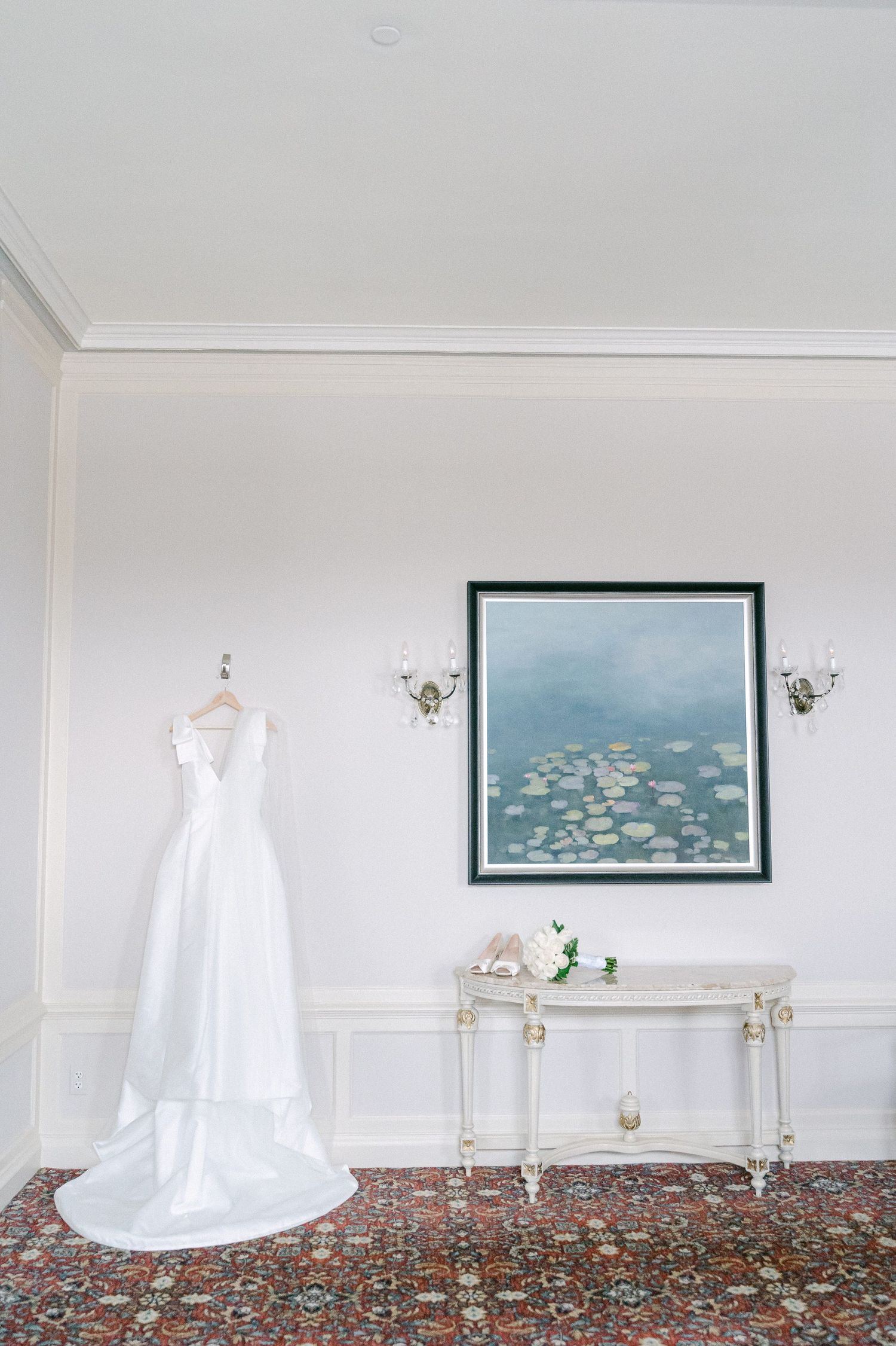 Elegant white wedding dress hanging on wall beside ornate console table with floral art and candle sconces in Philadelphia, PA