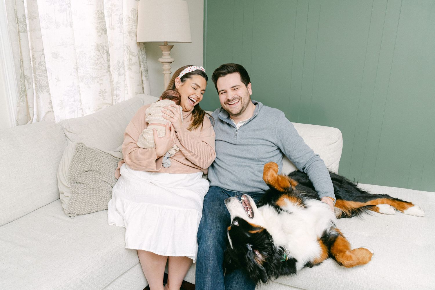A couple sits on a white couch with their Bernese Mountain Dog in a mint green room.
