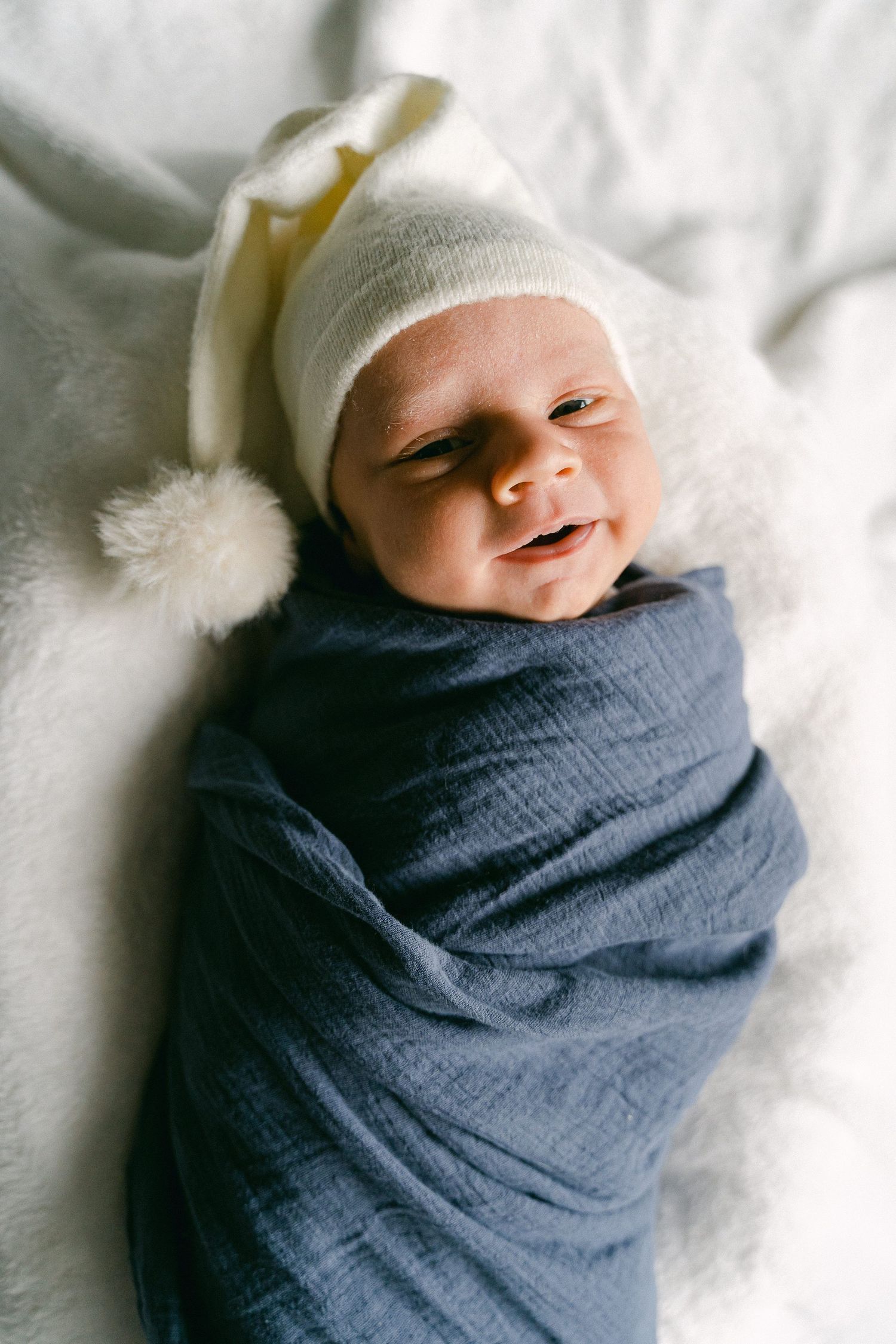A newborn baby swaddled in navy blue and white blankets smiles while resting on soft white bedding.