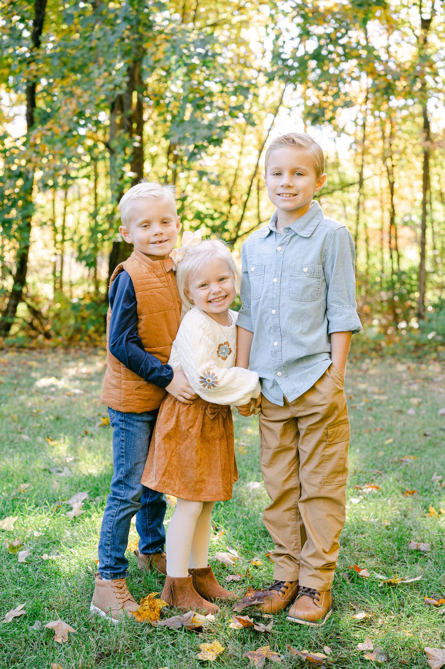 Three children in coordinating fall outfits stand together in a sunlit wooded area during an autumn photoshoot.