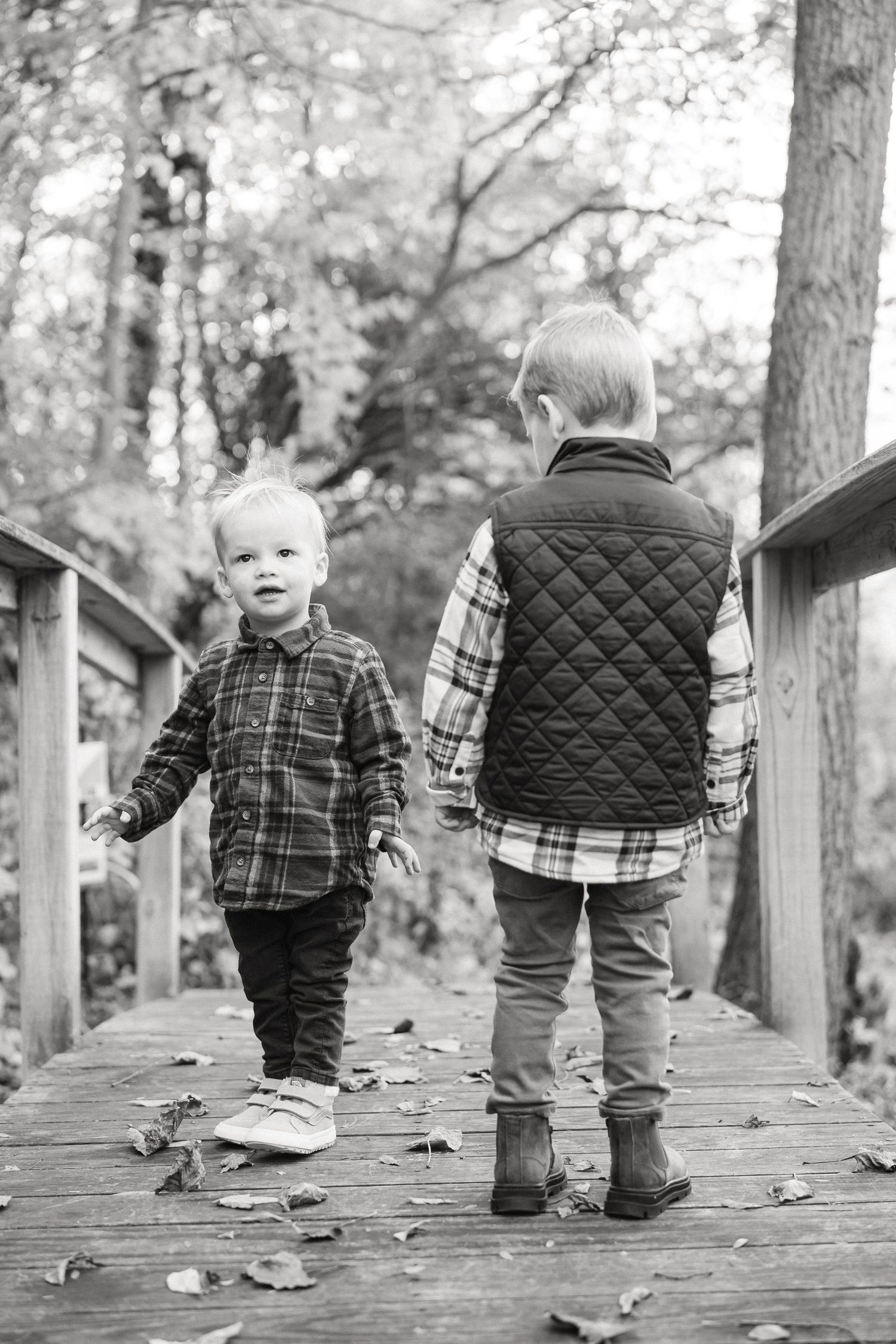 Black and white photo of toddler feet walking on wooden boardwalk with fallen leaves.