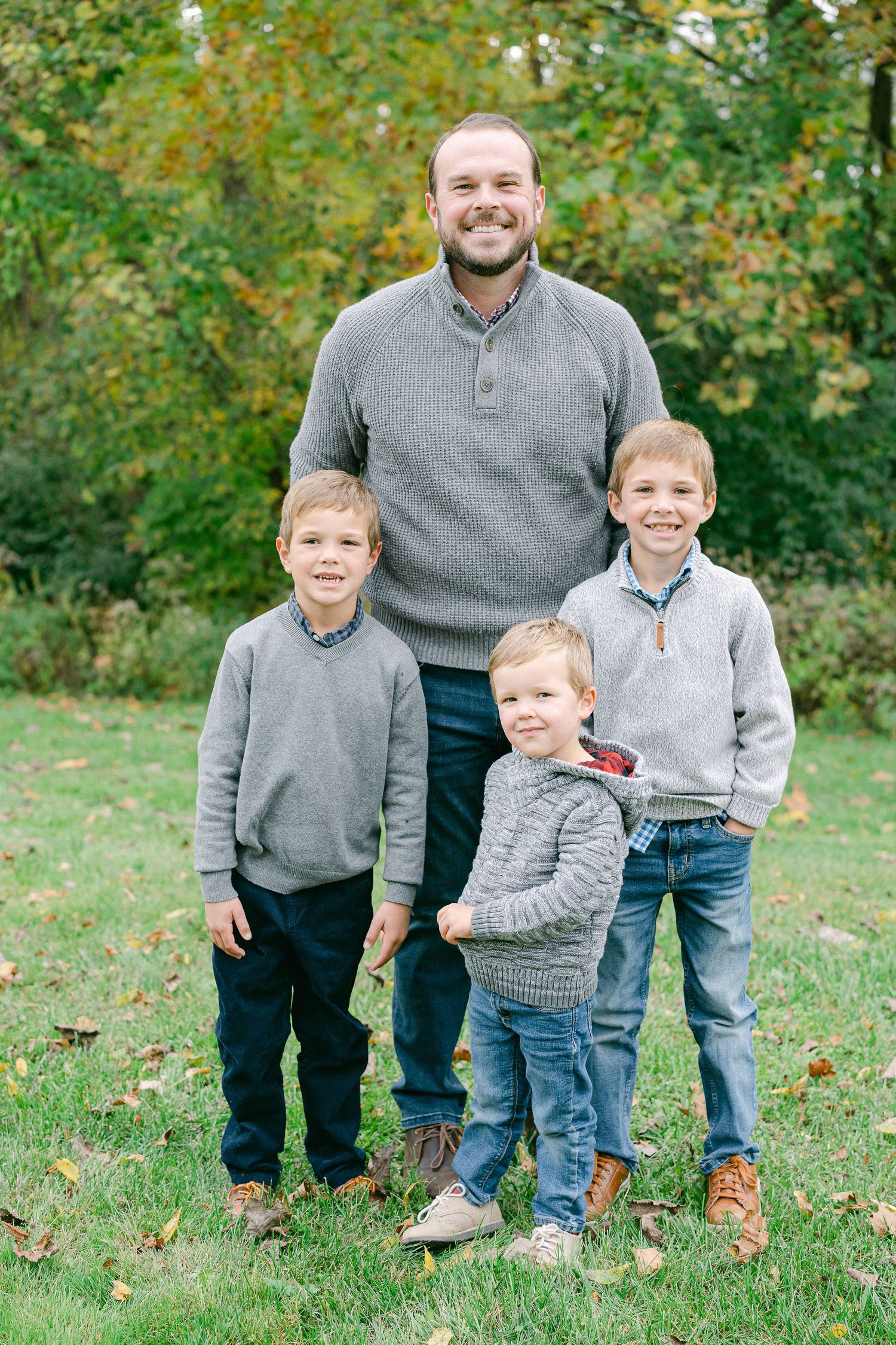 Family group wearing coordinating grey sweaters poses together on grass with autumn foliage in background.