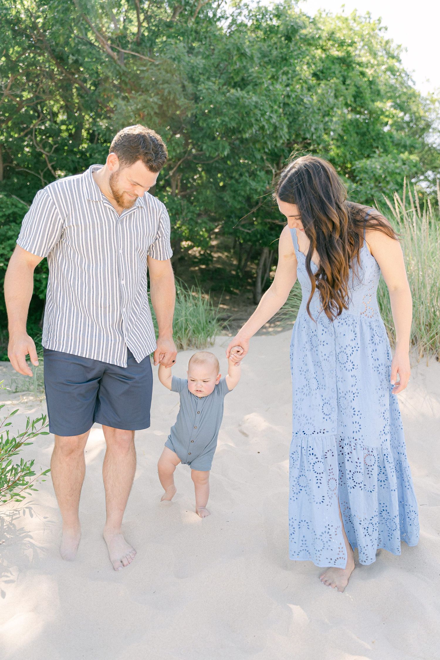 A couple walks barefoot together on a sandy path through greenery on a sunny day.
