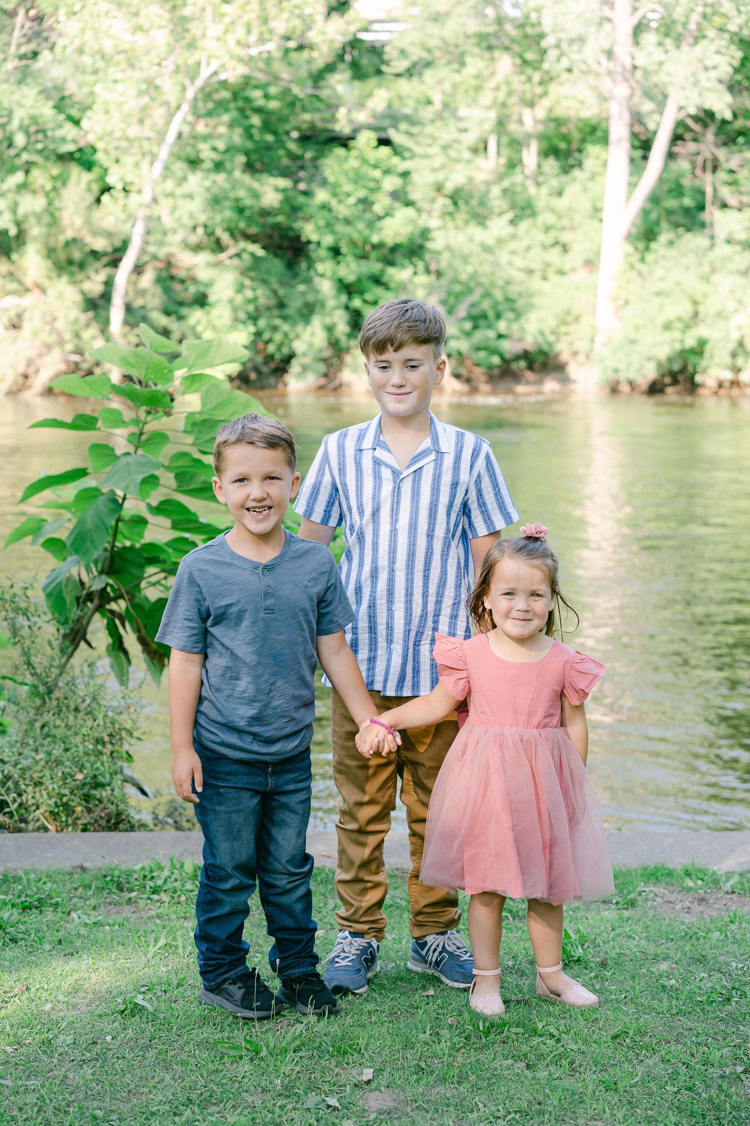 Three children stand together holding hands by a riverbank on a sunny day.