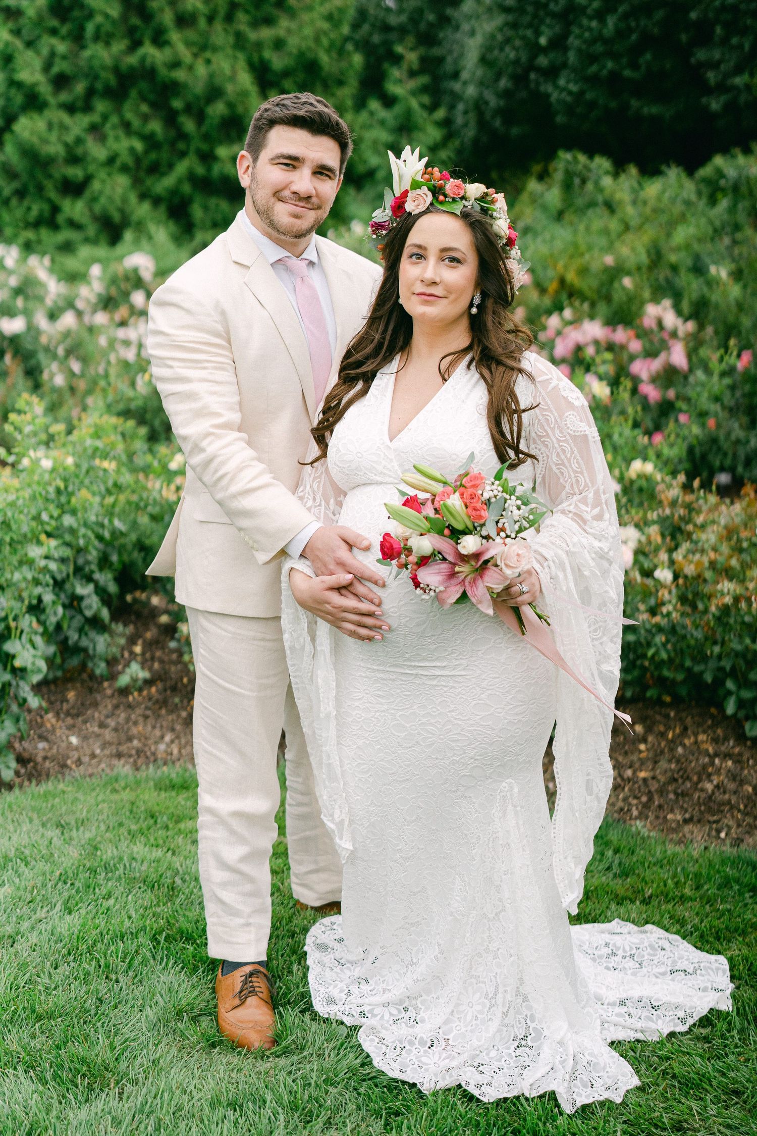 A romantic wedding couple poses together in a garden with pink flowers and lush greenery.
