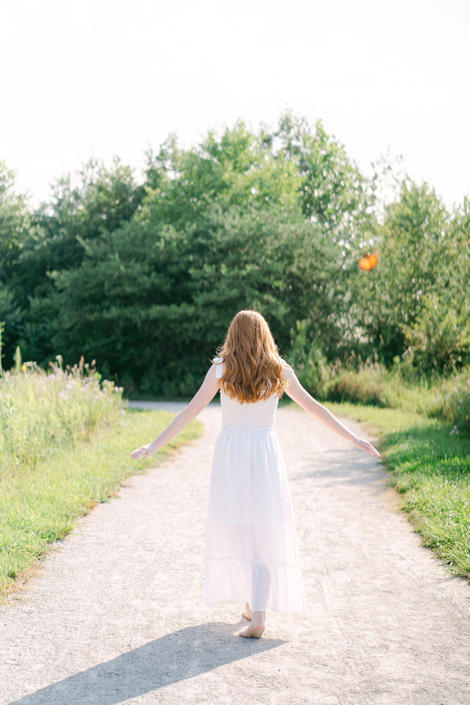 A figure in a flowing white dress twirls and dances along a sunlit nature path surrounded by lush greenery.
